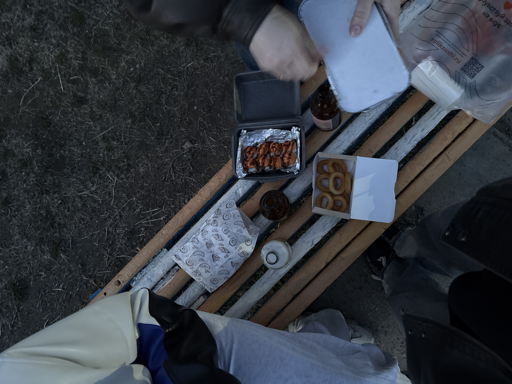 The image captures an outdoor scene on a slatted wooden bench. Several food items are arranged on the bench including a box of sushi, a box of onion rings, sauces, and a food container. Two individuals are visible, with one appearing to be opening the food container. The background consists of grassy terrain. The image suggests a casual outdoor meal setting. The location is unspecified and cannot be determined from the given image.
