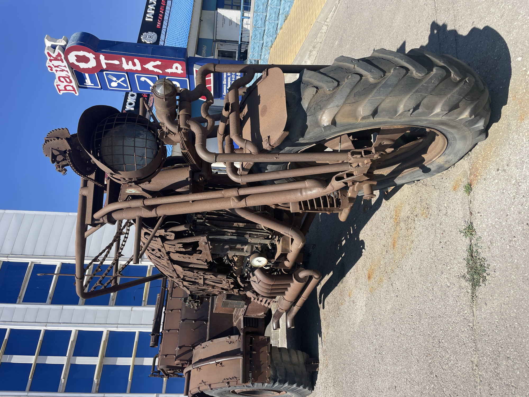 The image shows a large, rusted metal sculpture of a motorcycle parked on an asphalt surface in the city of Kamensk-Shakhtinsky, Russia. The sculpture is made of various metal parts, including pipes, gears, and what appears to be an engine. It has a large front wheel and a smaller rear wheel. In the background, there is a modern building with blue-tinted windows and a tall sign that reads "Байк ОТЕЛЬ" (Baik Hotel) in Cyrillic letters. There are no people visible in the image. The environment appears to be an outdoor area, possibly a parking lot or a public space. The sun is shining, casting shadows on the ground.