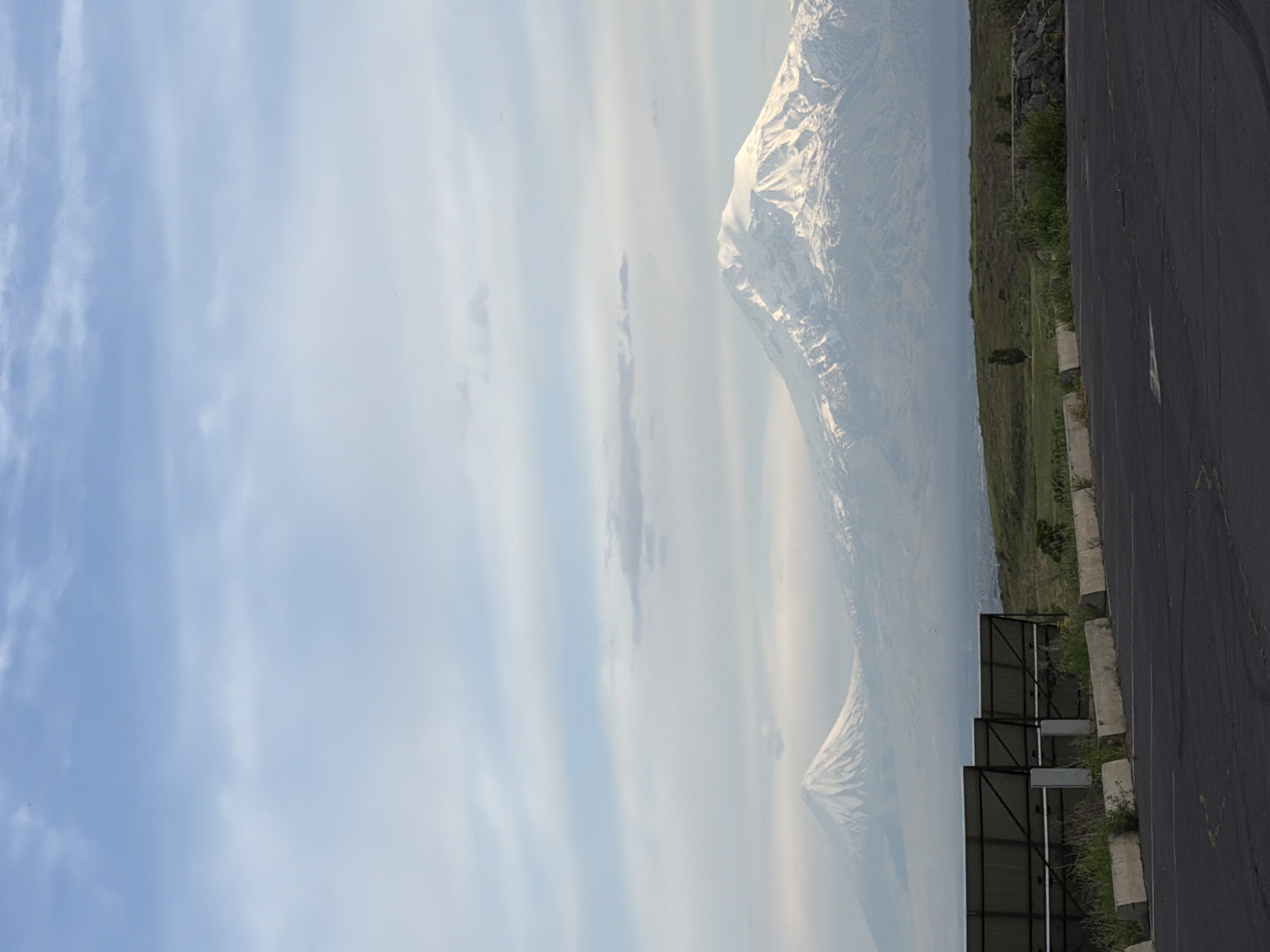The image depicts a view of Mount Ararat from Yerevan, Armenia. The mountain, partially covered in snow, is visible in the distance through a hazy atmosphere. In the foreground, there is a road with a line of solar panels along the side. The sky is light blue with sparse clouds. Vegetation is seen along the road's edge.