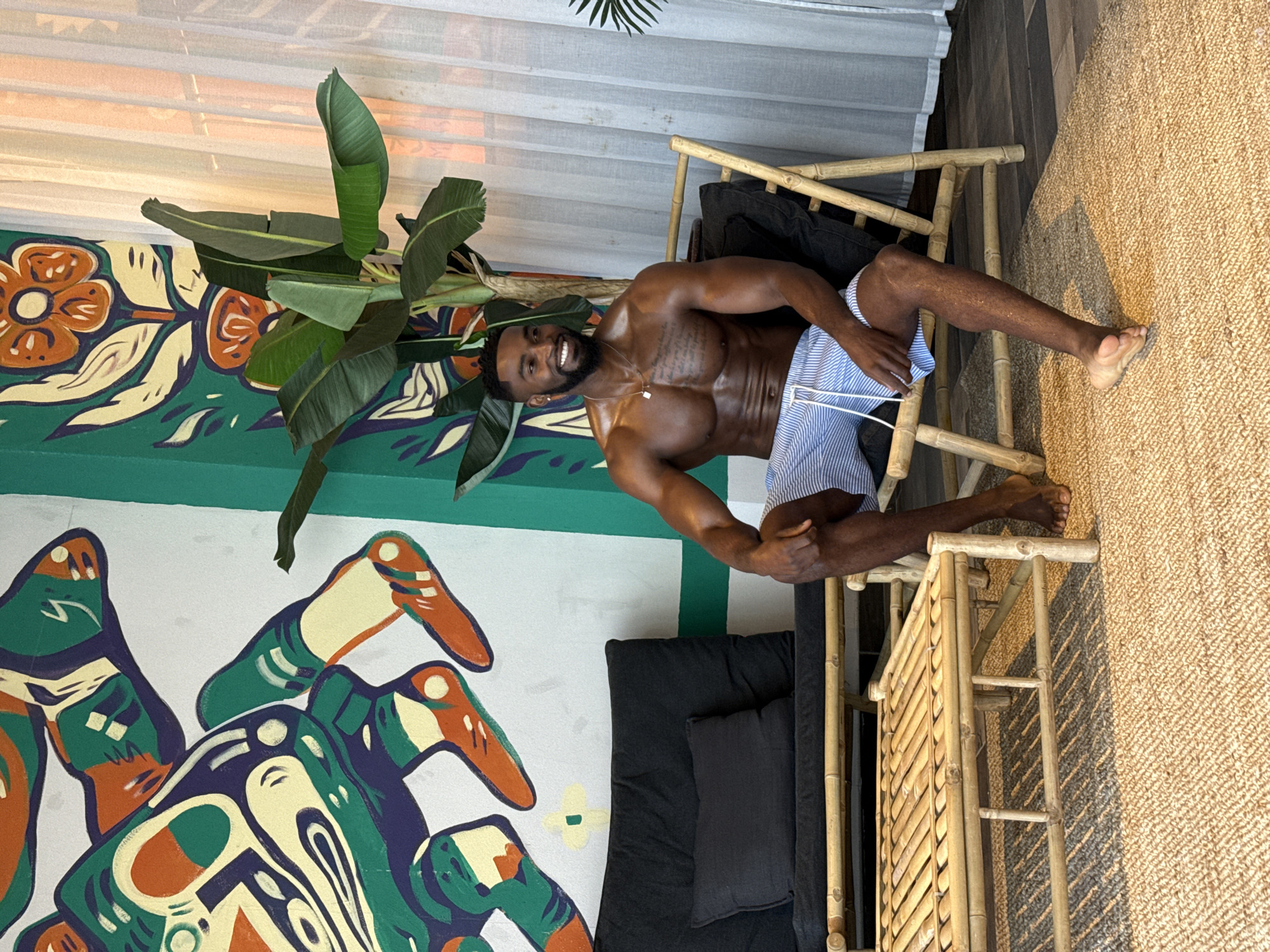 A smiling, muscular Black man is seated shirtless on a bamboo lounge chair with black cushions. He has a short beard and is wearing blue and white striped shorts. His body is angled slightly, with his head turned towards the viewer, conveying a relaxed and happy demeanor.
The interior setting features a vibrant, eclectic aesthetic. Behind the man, a wall is adorned with a colorful mural. The upper portion displays a teal-green background with abstract floral patterns in orange, cream, and dark blue. Below this, a larger, abstract design, possibly depicting a stylized insect or creature, utilizes a similar palette on a lighter background. A large green-leafed plant, resembling a banana plant, is positioned beside the man, adding a tropical touch.
The man's legs extend towards the right side of the frame, resting on a textured, woven rug, likely made of jute or sisal, which covers the floor and features a striped pattern. In the background, white sheer blinds cover a window, diffusing natural light into the space. The overall impression is a stylish and cheerful indoor environment, possibly a themed room in a residence or establishment in Levallois-Perret, France. No visible text is present.