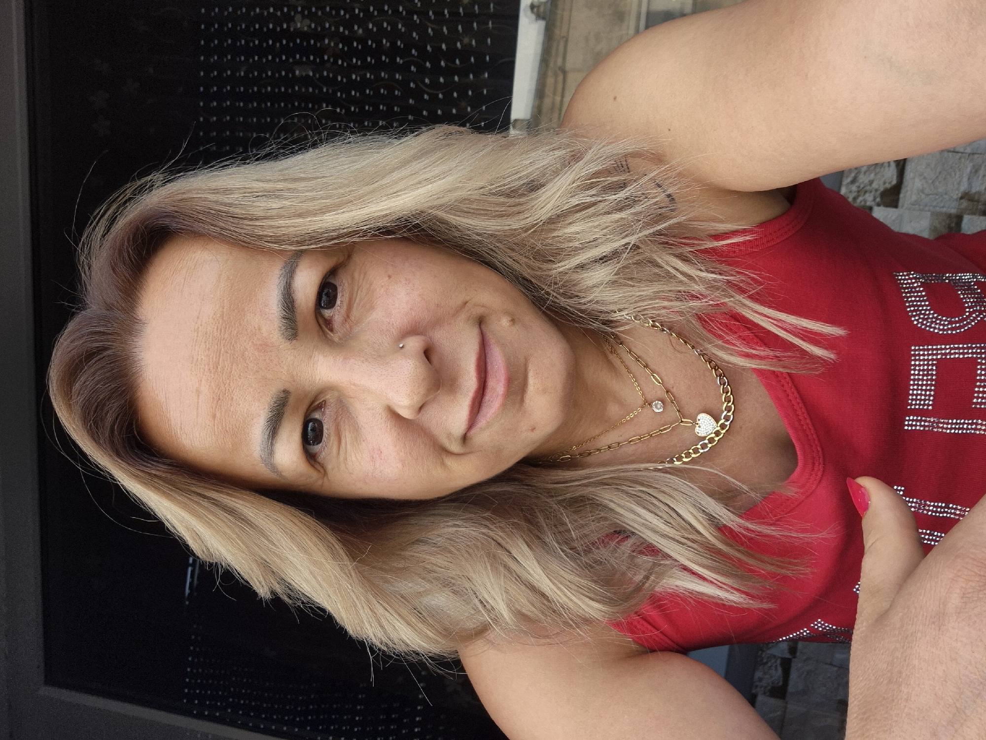 A medium shot captures a woman from the chest up, positioned outdoors. She has medium-length blonde hair, styled with waves, and her skin has a light complexion with some texture. The woman is looking directly at the camera with a slight smile. She is wearing a red tank top adorned with what appear to be rhinestones that form a pattern. Around her neck, she wears two gold necklaces, one with a small, clear pendant and the other with a heart-shaped pendant covered in small, clear stones. A subtle nose stud is visible on her right nostril. Her left arm is bent, and her hand is resting on her chest, with a bright pink fingernail visible. The background is slightly blurred, showing a dark, possibly windowed structure behind her on the left, and a textured wall or building facade on the right, suggesting an urban or semi-urban outdoor setting.