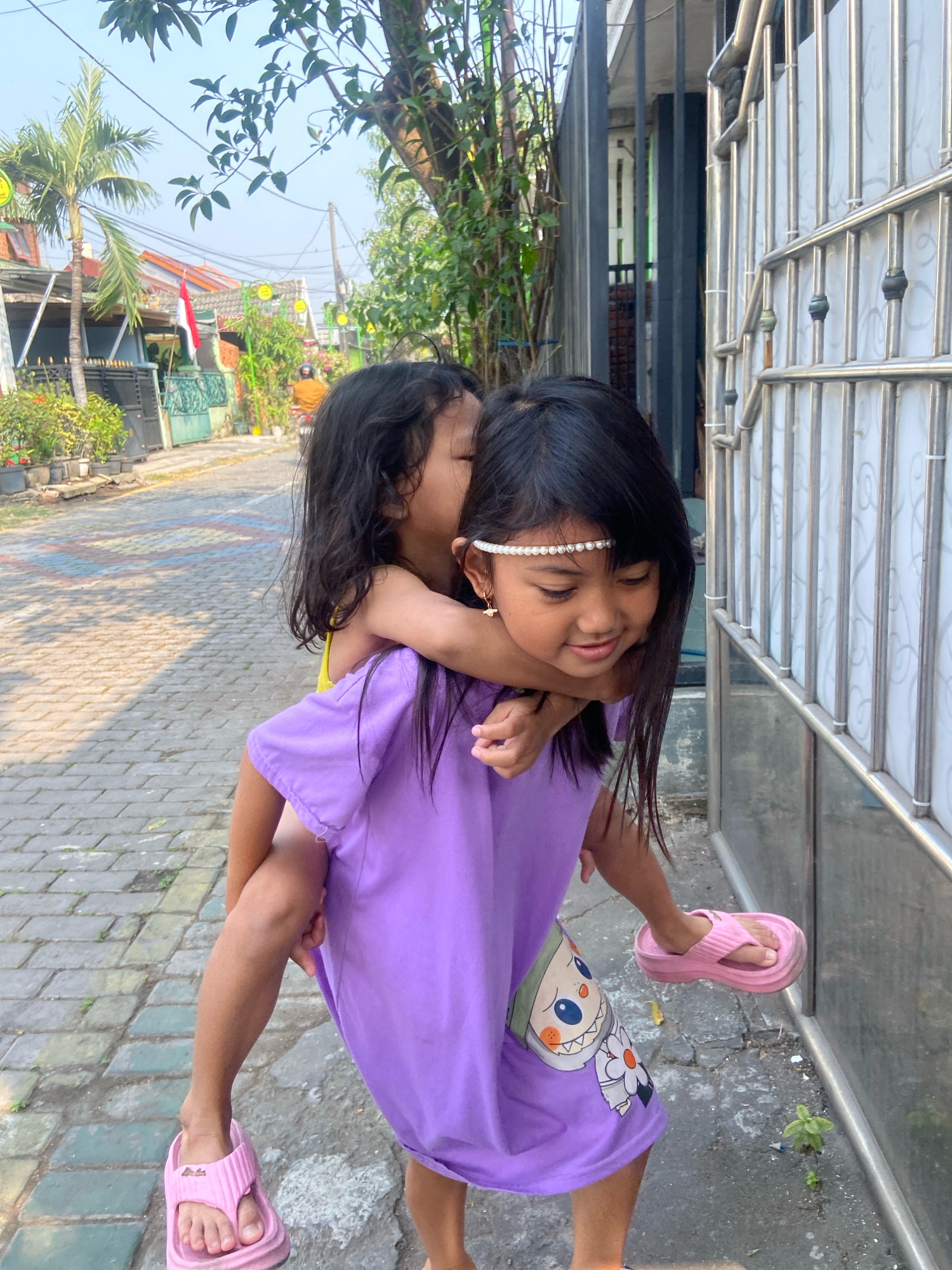 This image depicts two young girls in Gelam, Indonesia. The girl in the foreground, carrying the other on her back, is wearing a purple t-shirt with a cartoon character graphic and pink flip-flops. She has a pearl headband and small earrings. The girl on her back is wearing a yellow top and has her arms around the other girl's neck, whispering in her ear. They are standing on a paved walkway next to a metal fence. The background shows a street lined with houses, trees, and potted plants. The lighting suggests it is daytime, with shadows cast on the ground.