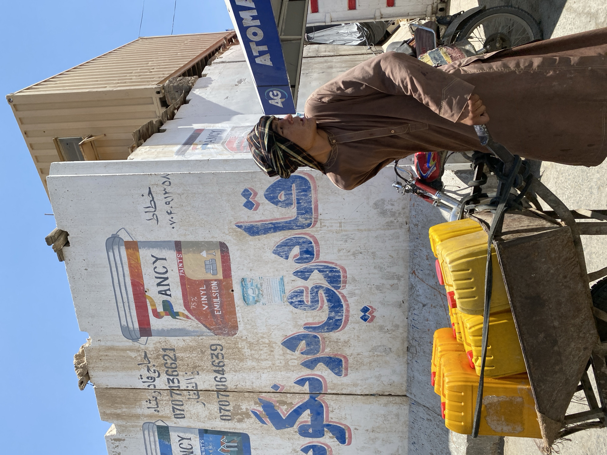 A young man, likely of Pashtun ethnicity and dressed in a traditional brown shalwar kameez and a checkered turban, stands next to a rustic wooden cart piled with several bright yellow plastic jerrycans. He is looking to his right, away from the camera. Behind him is a large concrete barrier, painted white, featuring advertising for "ANCY PAINTS" with images of paint cans, and Arabic script. The paint is peeling in places, revealing the concrete underneath. Further to the right, a blue awning with the text "ATOMA" and a "4G" logo is visible, suggesting a business or service. A red motorcycle is parked behind the cart and the man. The background shows a beige shipping container and more concrete structures, under a clear blue sky. The scene appears to be on a street in Kandahar, Afghanistan, with the concrete barrier likely part of security fortifications common in the region. The overall impression is one of everyday commerce or logistics taking place in a city environment marked by security measures.