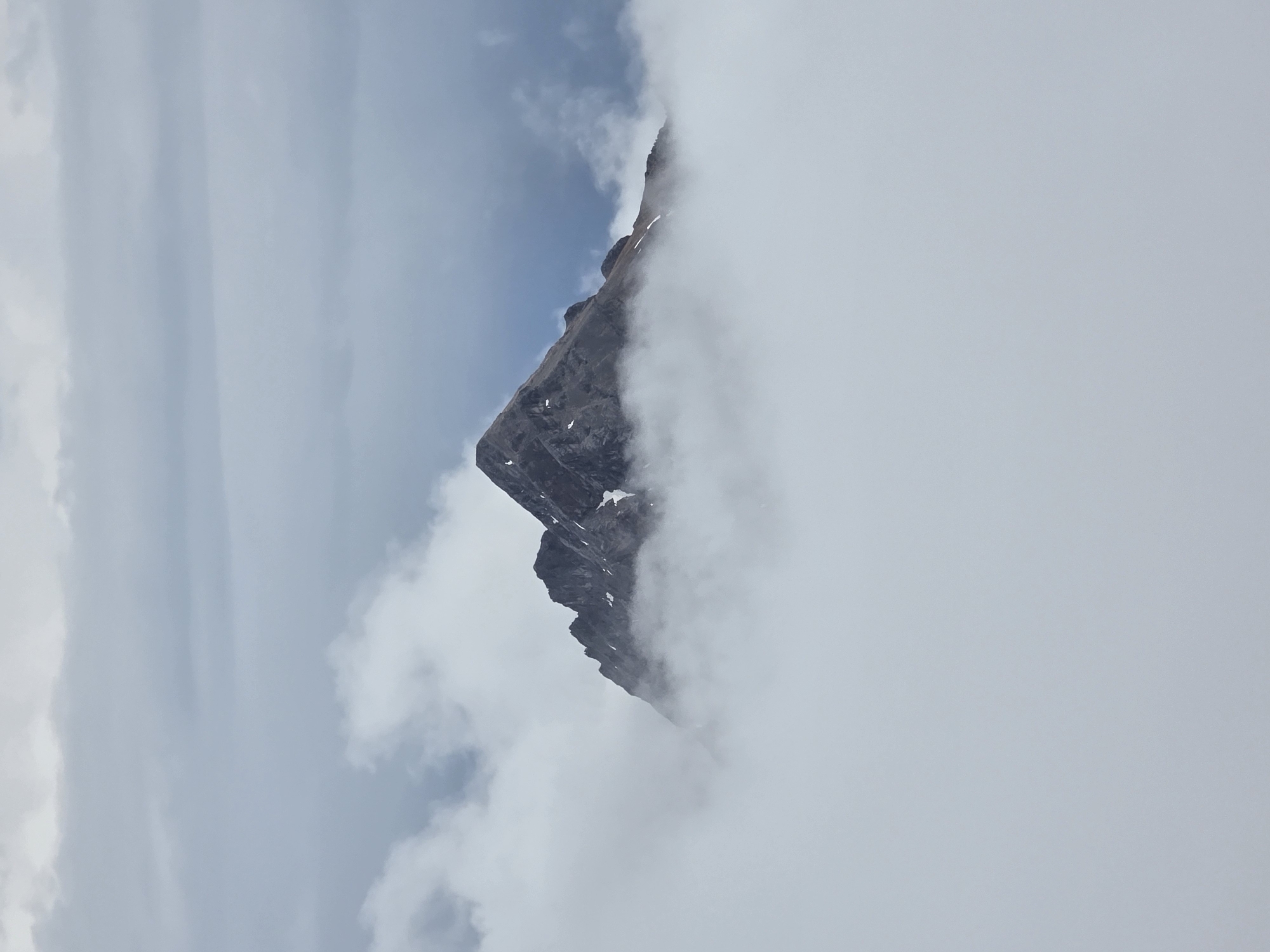 The image depicts a solitary mountain peak emerging from a thick blanket of clouds. The mountain is rugged and rocky, with some patches of snow visible on its slopes. The sky above is overcast with layered clouds, giving the scene a dramatic and somewhat somber atmosphere. The overall impression is one of isolation and the raw beauty of nature. The location is the Zugspitze, the highest peak in Germany, situated in the Wetterstein Alps near Garmisch-Partenkirchen.