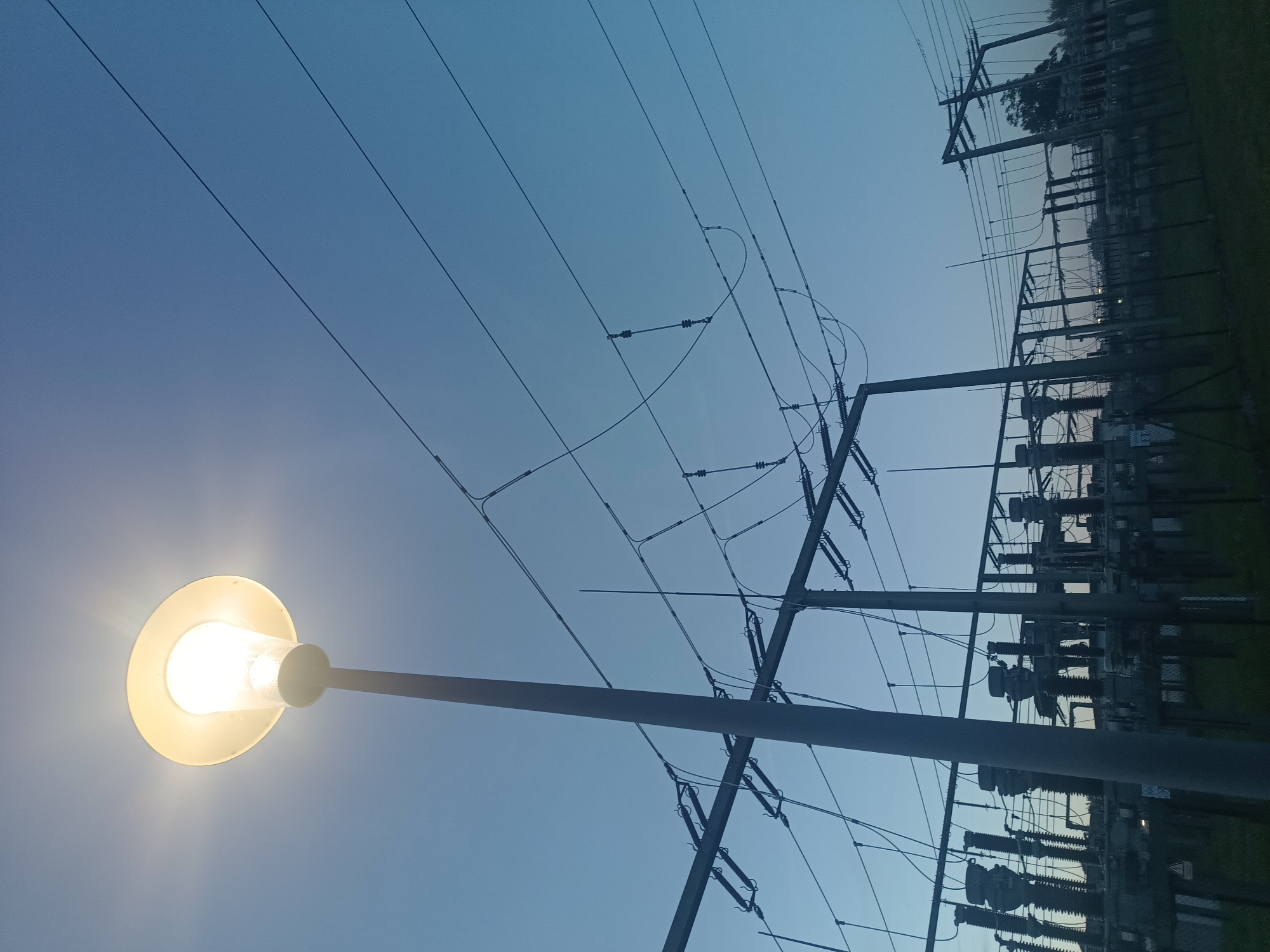 The image shows a streetlamp illuminating the sky over an electrical substation at dusk in Neuötting, Germany. The sky is a gradient of deep blue to a lighter blue, with the streetlamp emitting a bright yellow light from its frosted globe. A tall, dark gray pole supports the streetlamp. In the background, the substation is a complex of metal structures, insulators, and numerous overhead power lines. These lines are suspended from tall poles and stretch across the frame. The lighting suggests it is either dawn or dusk. The ground appears to be covered in grass, with some fencing visible. The overall scene conveys a sense of industrial infrastructure against a natural sky at twilight.