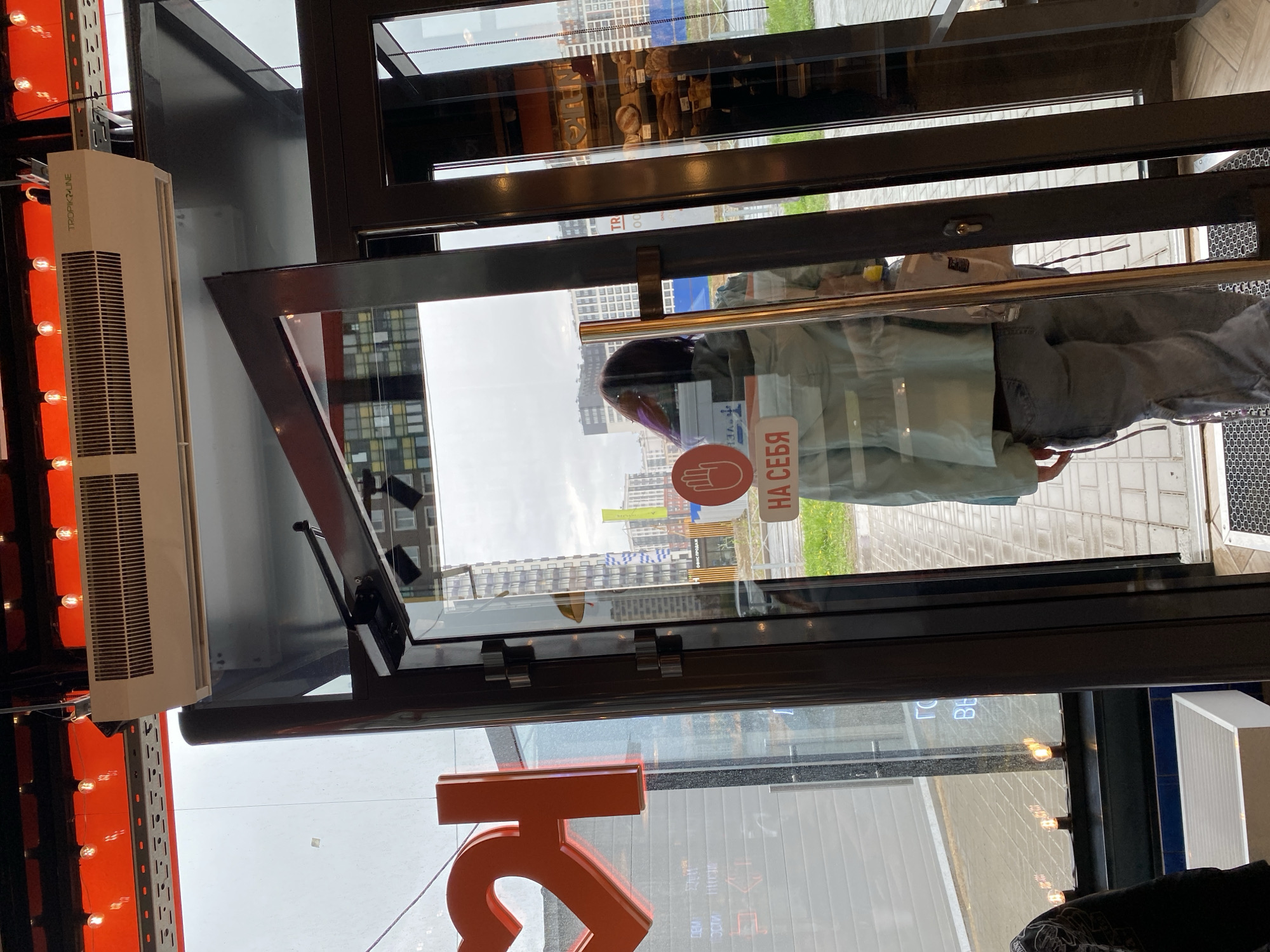 The photograph shows the interior of a building, likely a bakery or a similar food establishment, as suggested by the pastries visible in the glass display towards the top of the frame. Through the open glass door, an individual is seen entering the premises, their head with purple-streaked hair and torso partially visible. A red handprint sticker with Cyrillic text is affixed to the glass door. A prominent orange logo is displayed at the bottom left of the frame, and a white air conditioning unit is mounted on the left side of the door. Outside the door, the urban environment includes buildings, greenery, and tiled walkways, indicating that the location is in Murino, Russia. The overall scene is captured from an eye-level perspective.