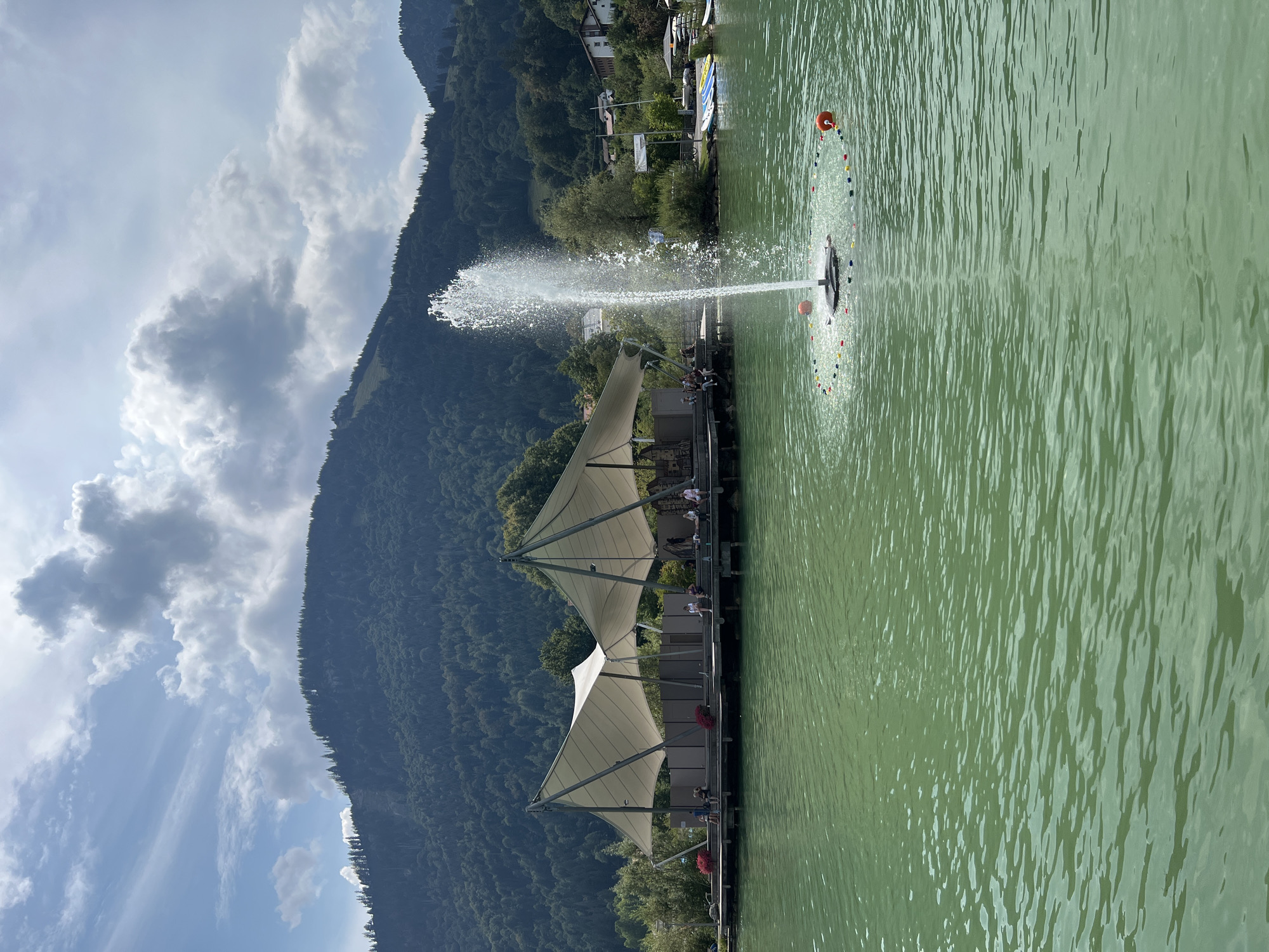 The image depicts a tranquil summer scene at the Alpsee in Immenstadt im Allgäu, Germany. In the foreground, a fountain vigorously sprays water into the lake, creating a refreshing spectacle. The lake itself is a vibrant green, with gentle ripples on its surface. A circular arrangement of small buoys surrounds the fountain.
In the mid-ground, a wooden pier juts out into the lake, supporting a stage with two large, sail-like awnings providing shade. Several people are seated on the pier, some appearing to be spectators, while others are casually conversing. Red flowers add pops of color to the edges of the pier.
The background is dominated by a lush, tree-covered mountain range, its slopes rising steeply towards a partly cloudy sky. The sky is a blend of blue and white, with dramatic clouds casting shadows and light on the landscape. Several buildings are visible nestled on the lower slopes of the mountain and along the lake's edge, suggesting a small town or village. To the right of the pier, several boats are docked, hinting at recreational activities on the lake. The overall atmosphere is one of peace and natural beauty.