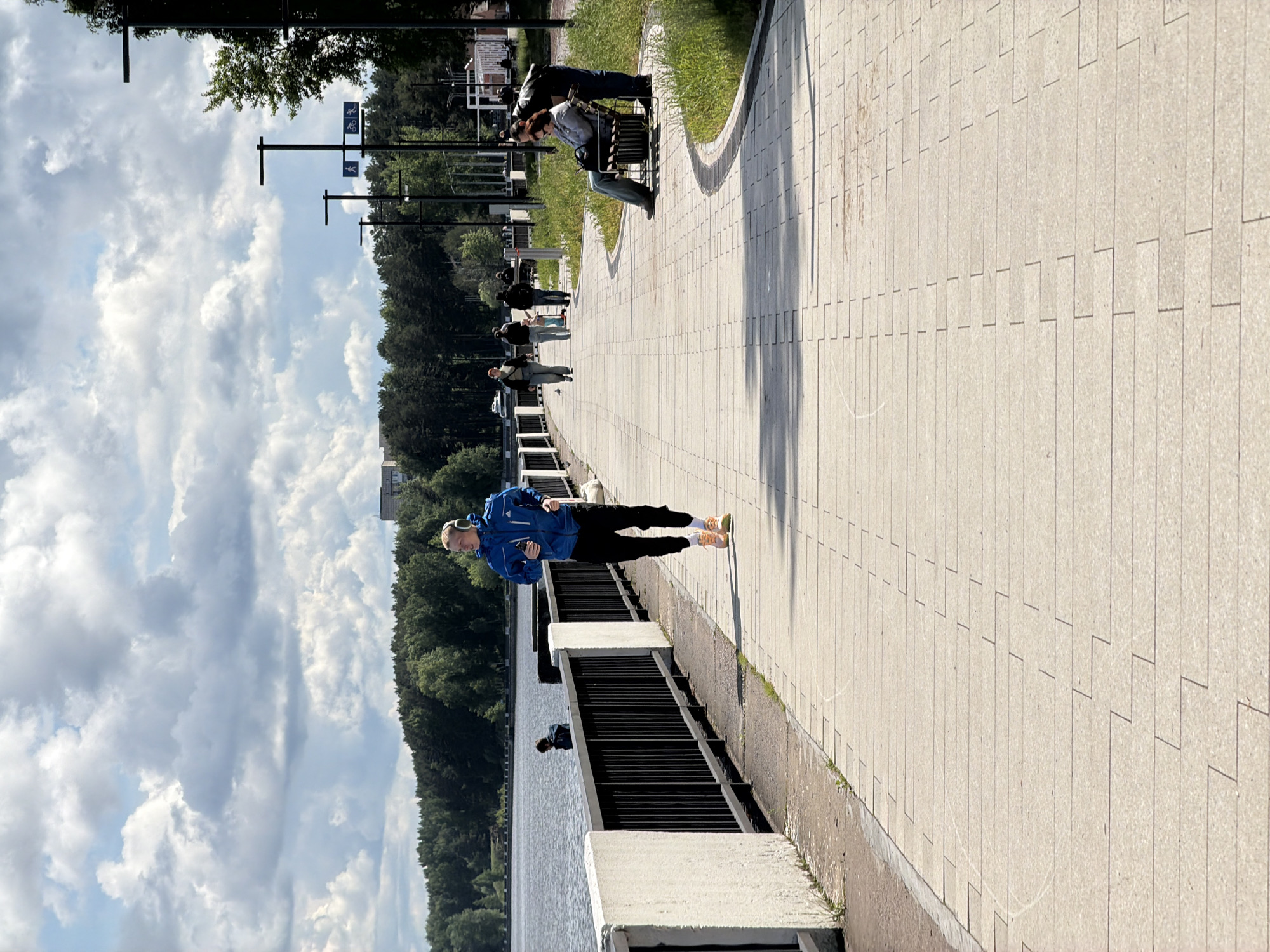 The image shows a man jogging on a paved path along a waterfront. He is wearing a blue jacket, black pants, and headphones. There are other people further down the path, including a couple sitting on a bench. The path is bordered by a black metal fence and then water. In the background, there are trees and a cloudy sky. This location is in the city of Chaikovsky, Russia.