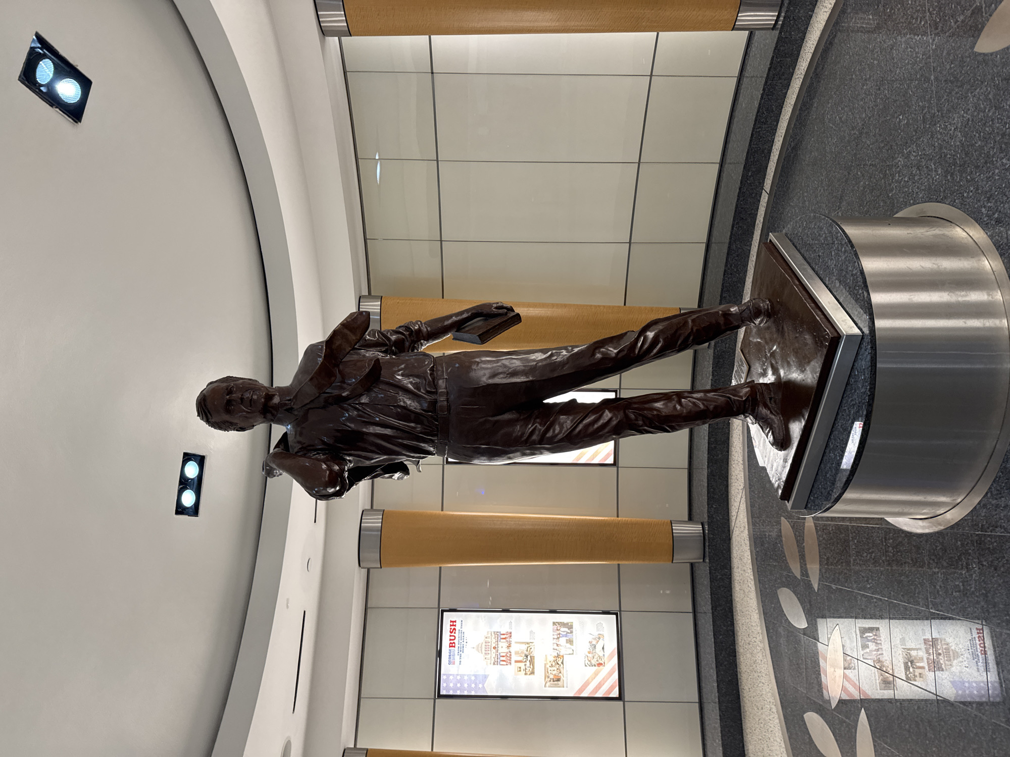 A full-body bronze statue of a man is positioned on a circular platform within an interior space. The statue depicts a man wearing a suit, walking, and holding a book. The statue is the main subject of the image and appears to be the statue of George H.W. Bush. The statue is surrounded by a circular walkway, lined with wall displays featuring the name "Bush" and images of buildings and documents. The flooring is dark and patterned with lighter-colored designs. The walls are composed of light-colored rectangular panels, accented by wooden cylindrical features with metal bands. Lighting fixtures are visible on the ceiling. Based on the presence of Bush's name and related imagery, the location is likely the George Bush Intercontinental Airport (IAH) in Houston, United States of America.