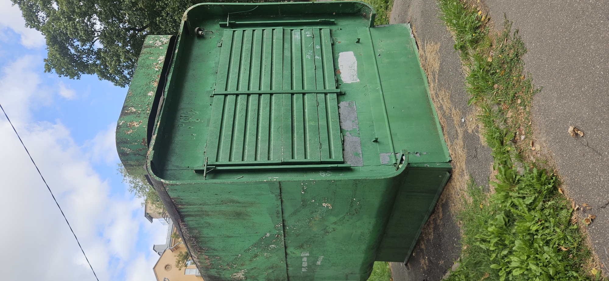 A weathered, dark green, metal kiosk stands on a paved surface bordered by grass and a sidewalk. The kiosk has a curved base and a flat top with a raised section. Its surface is extensively chipped and peeling, revealing rust and previous paint layers. There are two sets of horizontal, slatted shutters on the front of the kiosk, partially obscuring an opening. A thin black wire stretches across the upper part of the frame, against a partially cloudy sky. In the background, a building with a yellow facade and grey roof is visible, along with lush green trees. The ground around the kiosk is covered with patchy grass, dry leaves, and asphalt. The overall scene depicts an outdoor urban environment with a derelict-looking kiosk. The specific location in Vitsebsk, Belarus, is not discernible from the visual information provided.