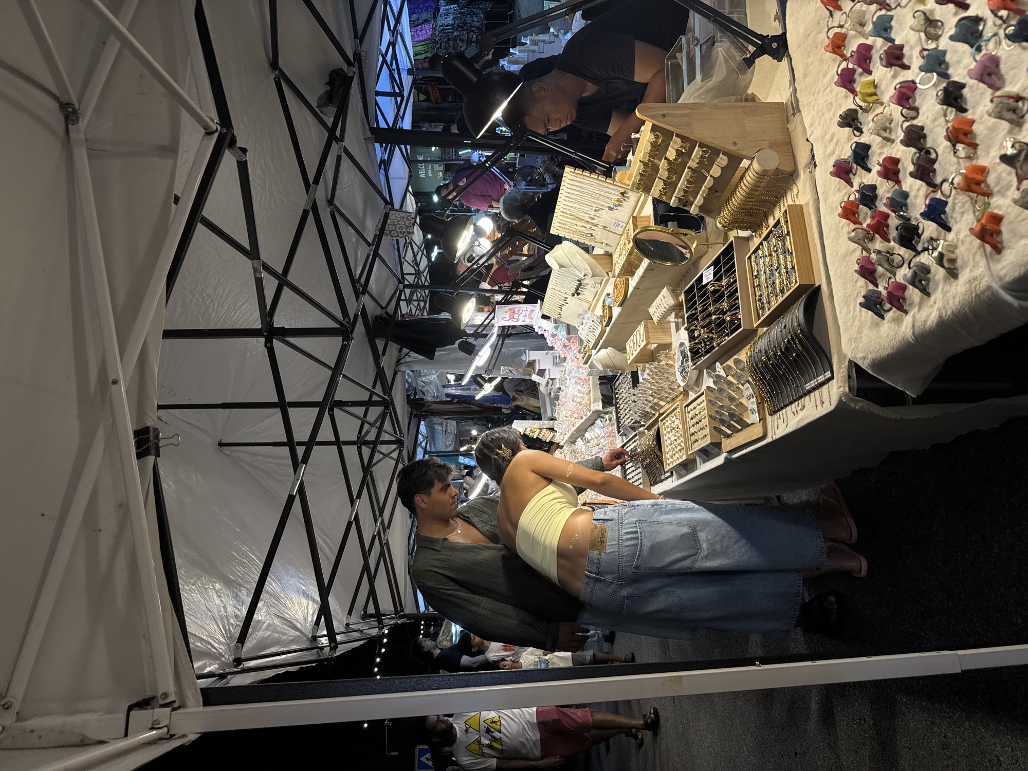 The image depicts a street market scene at night, likely in Phuket City Municipality, Thailand, given the presence of a stall selling jewelry and souvenirs. A couple is browsing a display of jewelry at a stall under a white canopy tent. The woman, wearing a light yellow crop top and wide-leg denim pants, leans over the table, while the man, in a dark green open-collar shirt, stands behind her, looking at the jewelry. The stall is laden with various necklaces, bracelets, earrings, and rings displayed on wooden stands and in trays. Several bright desk lamps illuminate the merchandise. In the background, other market-goers and stalls are visible, suggesting a bustling evening atmosphere. A man in a dark t-shirt is seated behind the jewelry display, seemingly attending to customers. The ground is paved asphalt.
