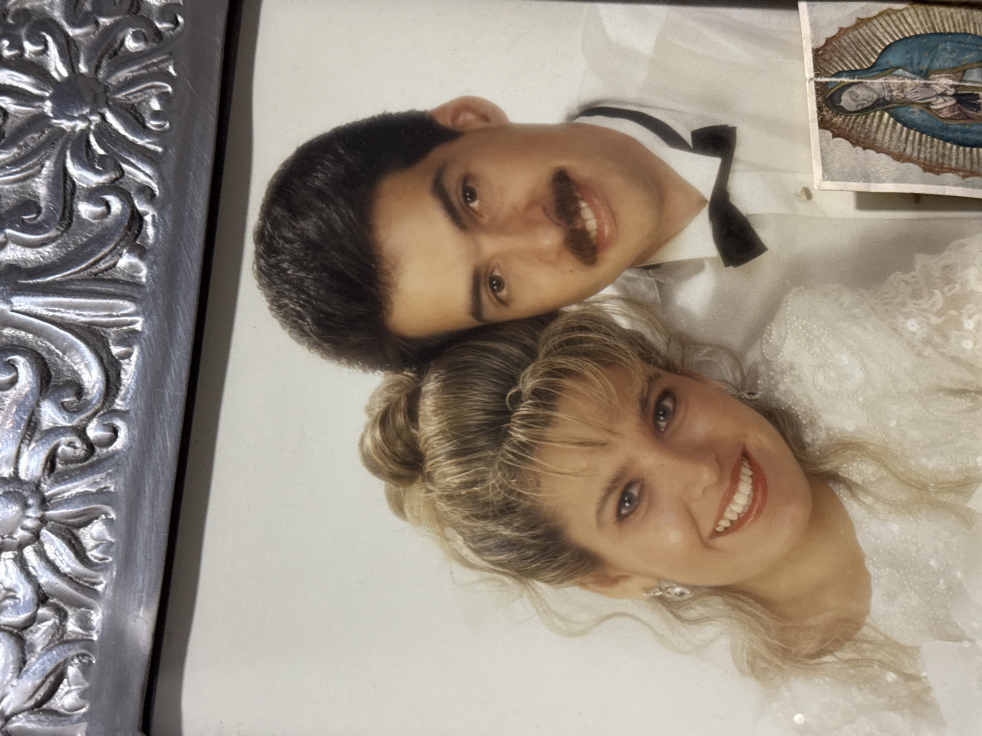 A framed wedding photograph shows a couple. The man has dark hair, a mustache, and wears a white shirt with a black bow tie. The woman has blonde hair and wears a white dress with a lace design. A small religious card depicting an image of the Virgin Mary is also visible in the top right corner of the frame. The frame itself is ornate and silver in color. The location of Santa Clara Ocoyucan, Mexico is not discernible from the image.