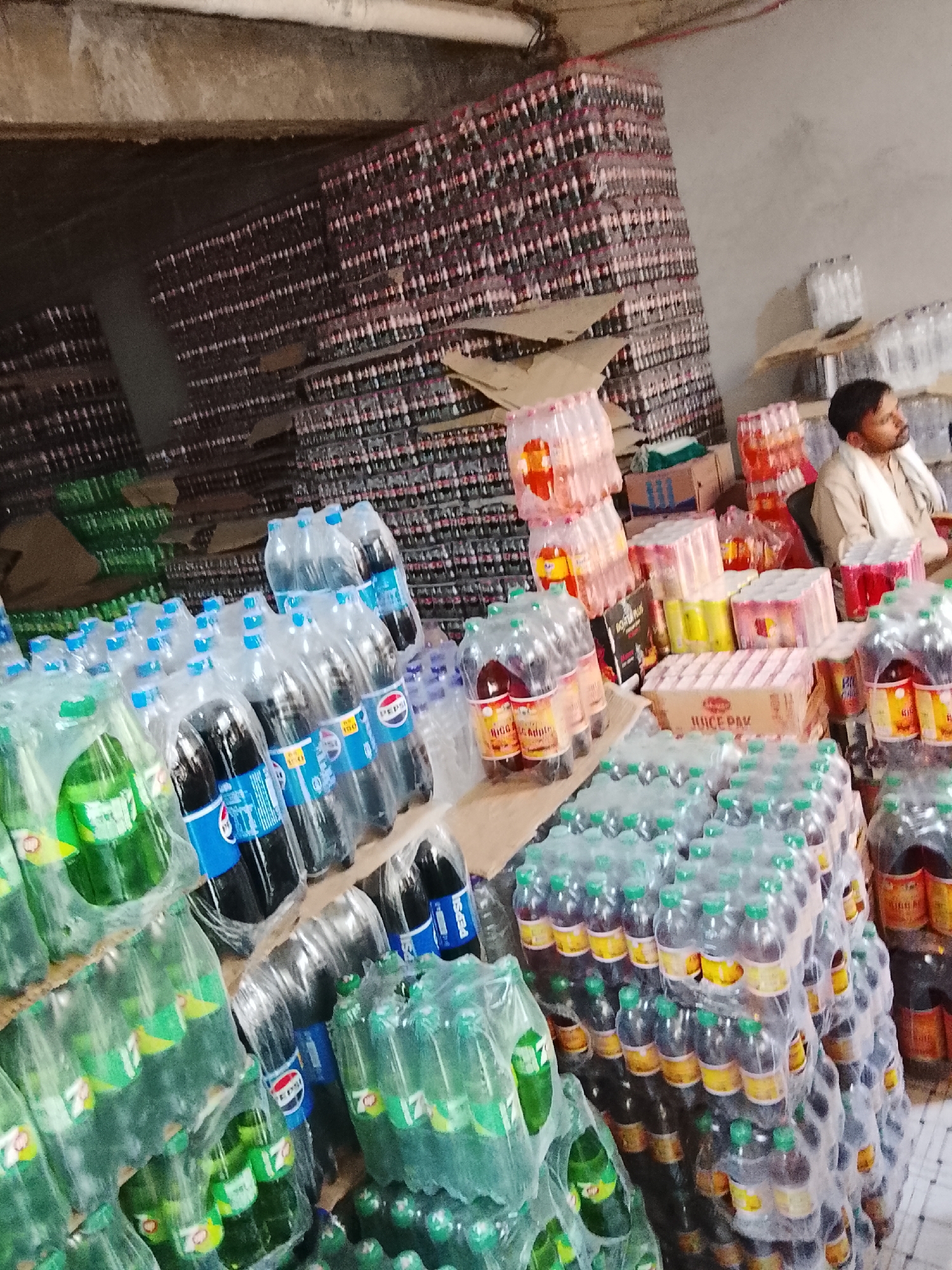 The image shows a warehouse or storage area in Gulgasht Colony, Pakistan. Stacks of bottled beverages in plastic wrap are the primary subjects. Brands like Pepsi and 7-Up are visible, along with other packaged juices and drinks. A man is seated in the background near shelves, seemingly observing the inventory. The location appears to be a large-scale distribution or storage facility given the volume of product.