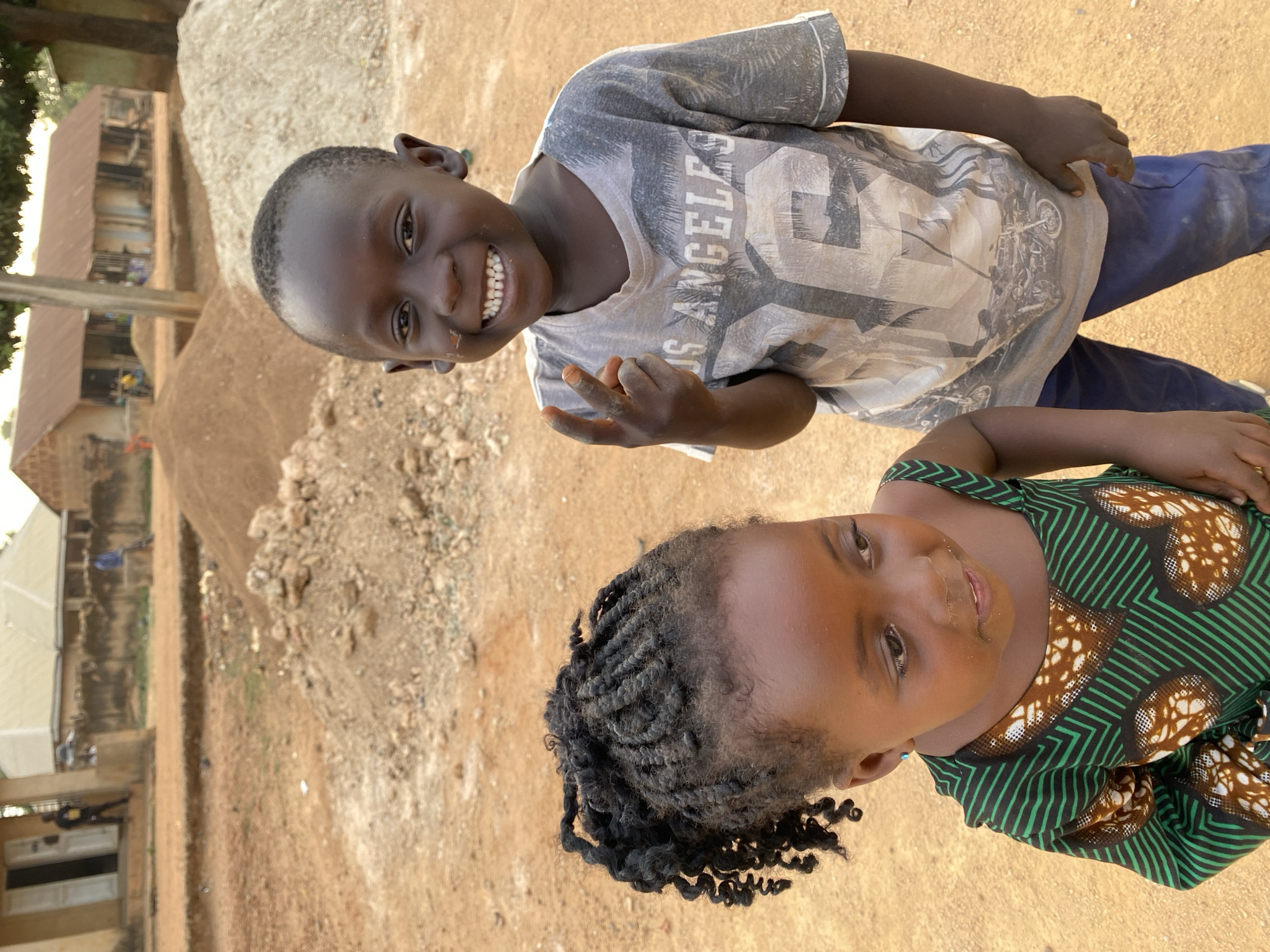 Two dark-skinned children, likely a boy and a girl, are posing for a photo outdoors in Kuru Dungus, Nigeria. The boy, on top, is wearing a grey t-shirt with "Los Angeles 96" printed on it and blue pants. He is smiling broadly, showing his teeth, and making a V sign with his fingers. The girl, below, is wearing a green and brown patterned dress. Her hair is styled in braids. The background shows a dusty, sandy ground with a pile of dirt or gravel. There are buildings visible in the distance, suggesting a residential or rural area.
