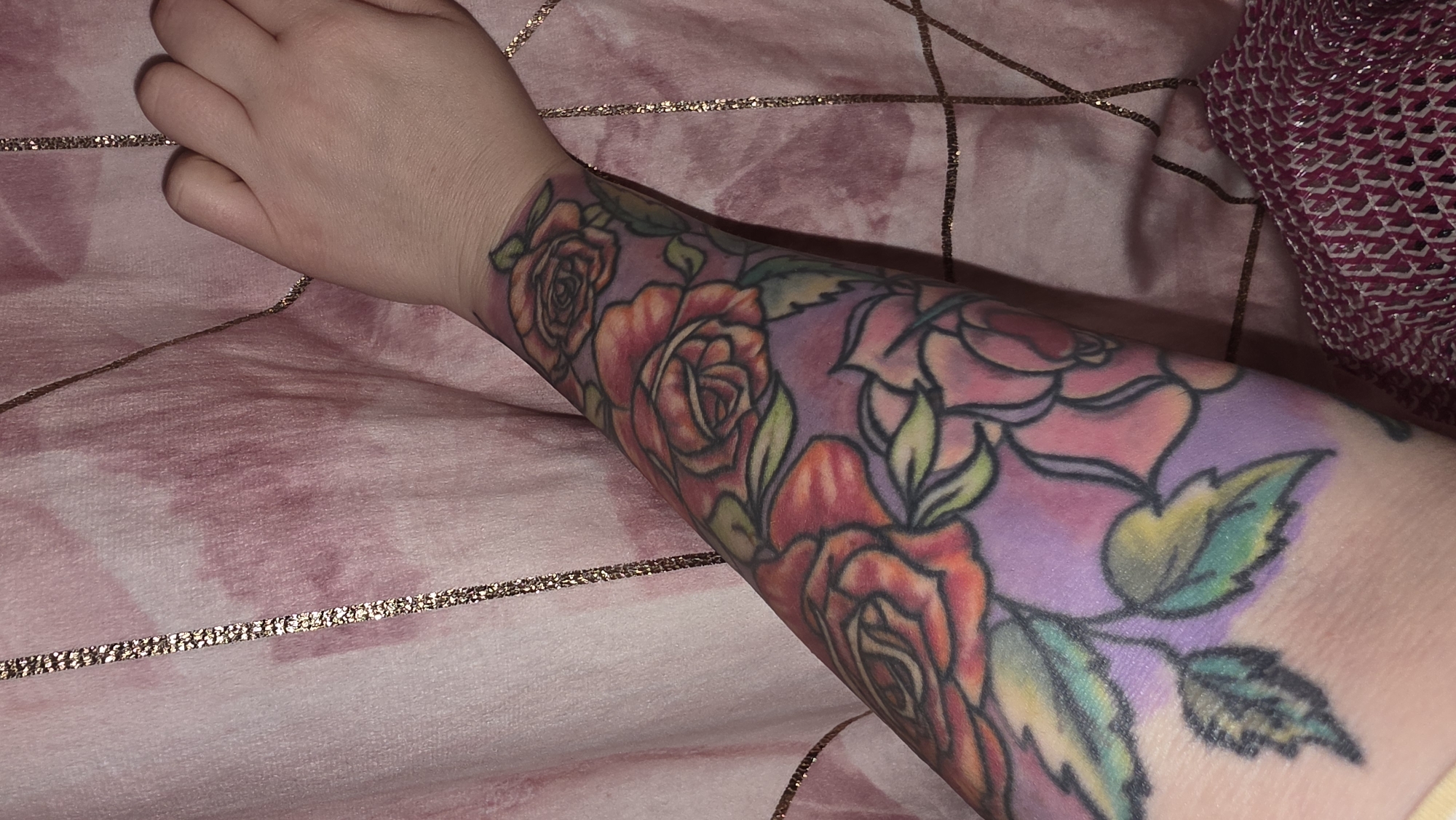 A close-up photograph shows a person's forearm with a colorful tattoo of roses and leaves, resting on a pink and white patterned fabric. The tattoo covers the entire forearm from wrist to elbow, featuring various roses in shades of red, orange, and pink, with green and yellow leaves. The background of the tattoo is a soft purple hue. The person's hand is visible at the top of the frame. The fabric underneath appears to be a bedspread or blanket with gold grid lines. Part of a textured pink fabric is visible to the right. There is no evidence to suggest it is in London.