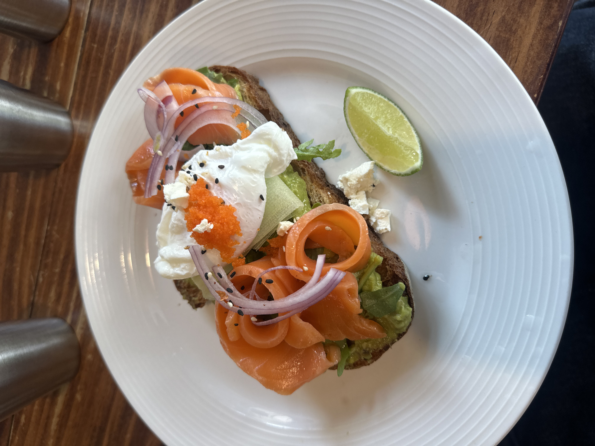 A high-angle, close-up shot reveals a plate of food on a wooden surface. The plate is white with concentric ring patterns. The food consists of toast topped with avocado, smoked salmon, poached eggs, red onion rings, fish roe, feta cheese, and a lime wedge. The location in the city of Đà Nẵng, Vietnam is not discernible from the image.