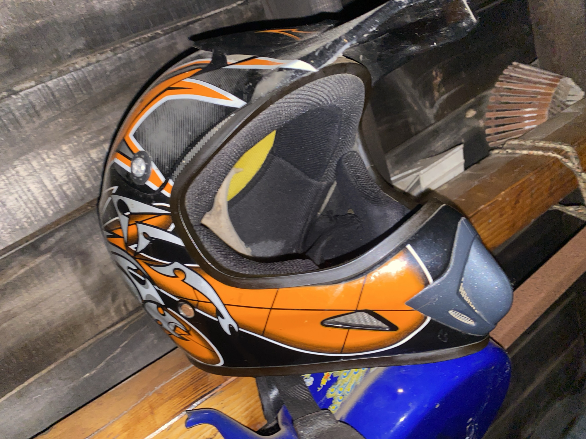 The image shows an orange, black, and silver motocross helmet resting on top of a blue plastic object. The helmet has a visor and interior padding visible. The objects are located indoors, against a background of wooden boards and other unidentified items. The location is Konstantinovo, Bulgaria.