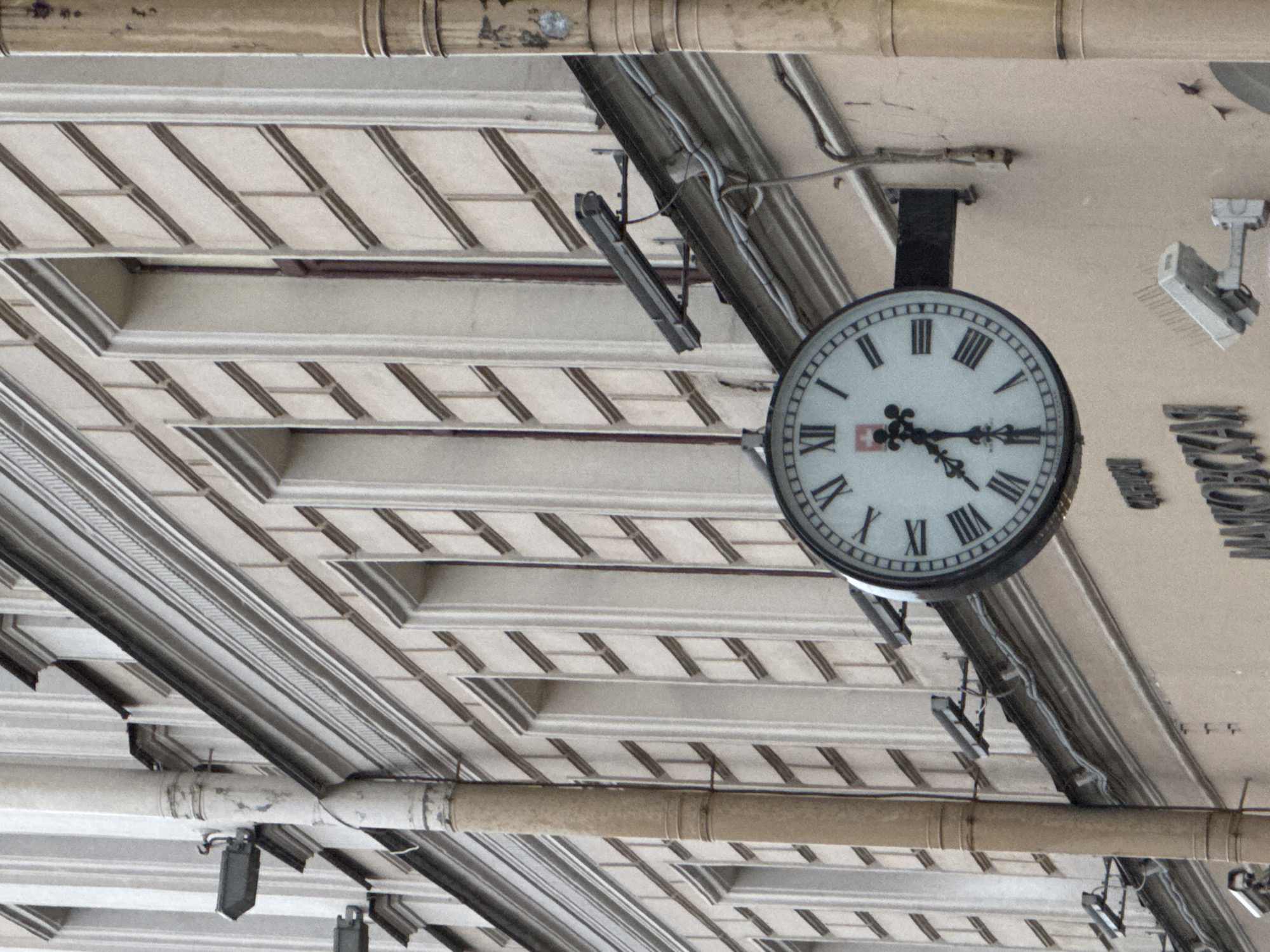 This image captures an upward view of a public indoor ceiling, likely from a train station or a large building in Saint Petersburg, Russia. The primary subject is a large, ornate double-sided clock hanging from a dark bracket. The clock face is white with black Roman numerals (I to XII) and has a black minute hand pointing to approximately 9 and an hour hand between 1 and 2, indicating the time is around 1:45 or 1:46. A small red and white emblem is visible near the VI numeral.
The ceiling features a repeating pattern of light-colored, recessed rectangular and square panels, suggesting a decorative and structured design. Running horizontally across the scene are several large, light-colored pipes, likely part of the building's infrastructure. Beneath these pipes and integrated into the ceiling structure are various electrical conduits, cables, and numerous light fixtures or security cameras mounted at different angles.
To the right of the clock, partially obscured and arranged vertically, is Cyrillic text that appears to spell out "МОСКОВСКИЙ" (Moskovskiy), which is consistent with the Moskovsky Railway Station in Saint Petersburg. No people are visible, and there is no specific activity taking place, depicting a static architectural detail. The lighting is even and soft, suggesting an indoor environment or an overcast day, with artificial light illuminating the space.