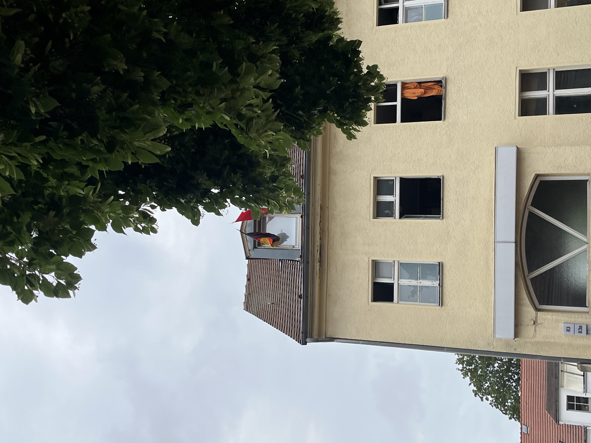 This image captures a close-up view of a yellow apartment building, partially obscured by the lush green foliage of a large tree. The building features multiple windows, some of which are open, revealing glimpses of the interior. One window on a higher floor displays an orange fabric. On a balcony, a German flag is visible, consisting of three horizontal stripes: black, red, and gold. The building's facade is textured, and architectural details like window frames and a large, semicircular window on the right are apparent. Below the main building, the red-tiled roof of another structure can be seen. The sky is overcast, suggesting a cloudy day. The scene appears to be in an urban residential area, possibly Berlin, Germany, given the German flag. No people are visible in the frame. The overall mood is neutral, with the focus on the architectural elements and the natural presence of the tree. Small numbers "83" and "83a" are visible on the wall near the semicircular window.