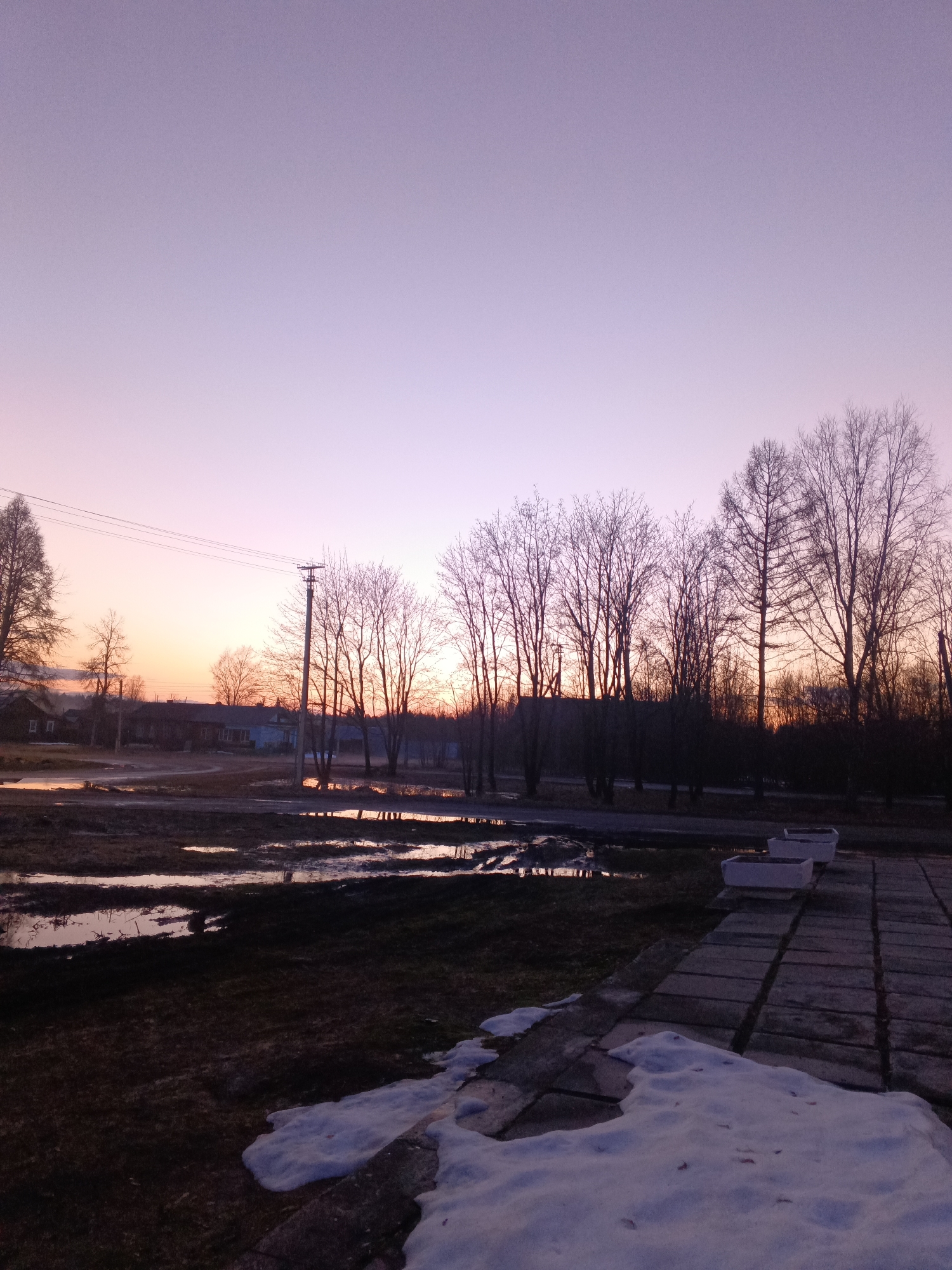 The image captures a twilight scene in Красный Маяк, Russia. Patches of melting snow sit on a paved area in the foreground. Beyond, puddles reflect the fading light across a field. Bare trees stand between the viewer and a row of houses in the middle ground. Power lines stretch across the sky. The sky exhibits a color gradient from orange on the horizon to purple above.