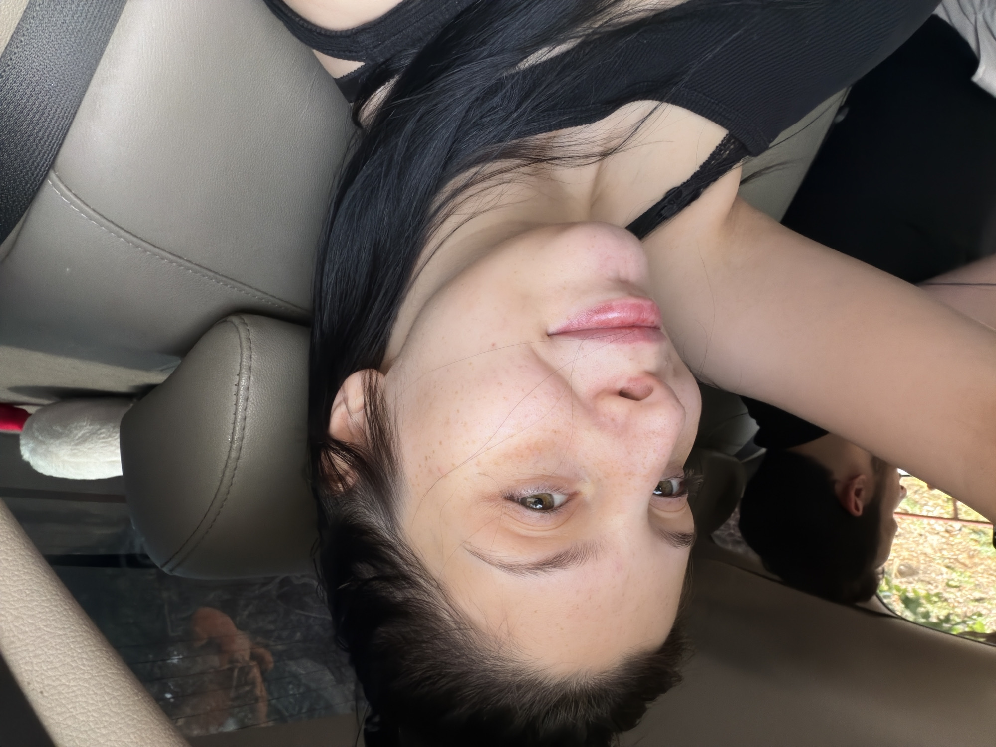 A young woman with black hair and freckles is in the passenger seat of a car, taking a selfie. She is wearing a black tank top. A man with dark hair is in the driver's seat, partially visible. The interior of the car appears to be a light beige leather. Outside the window, trees and foliage are visible. The image was taken during the daytime.