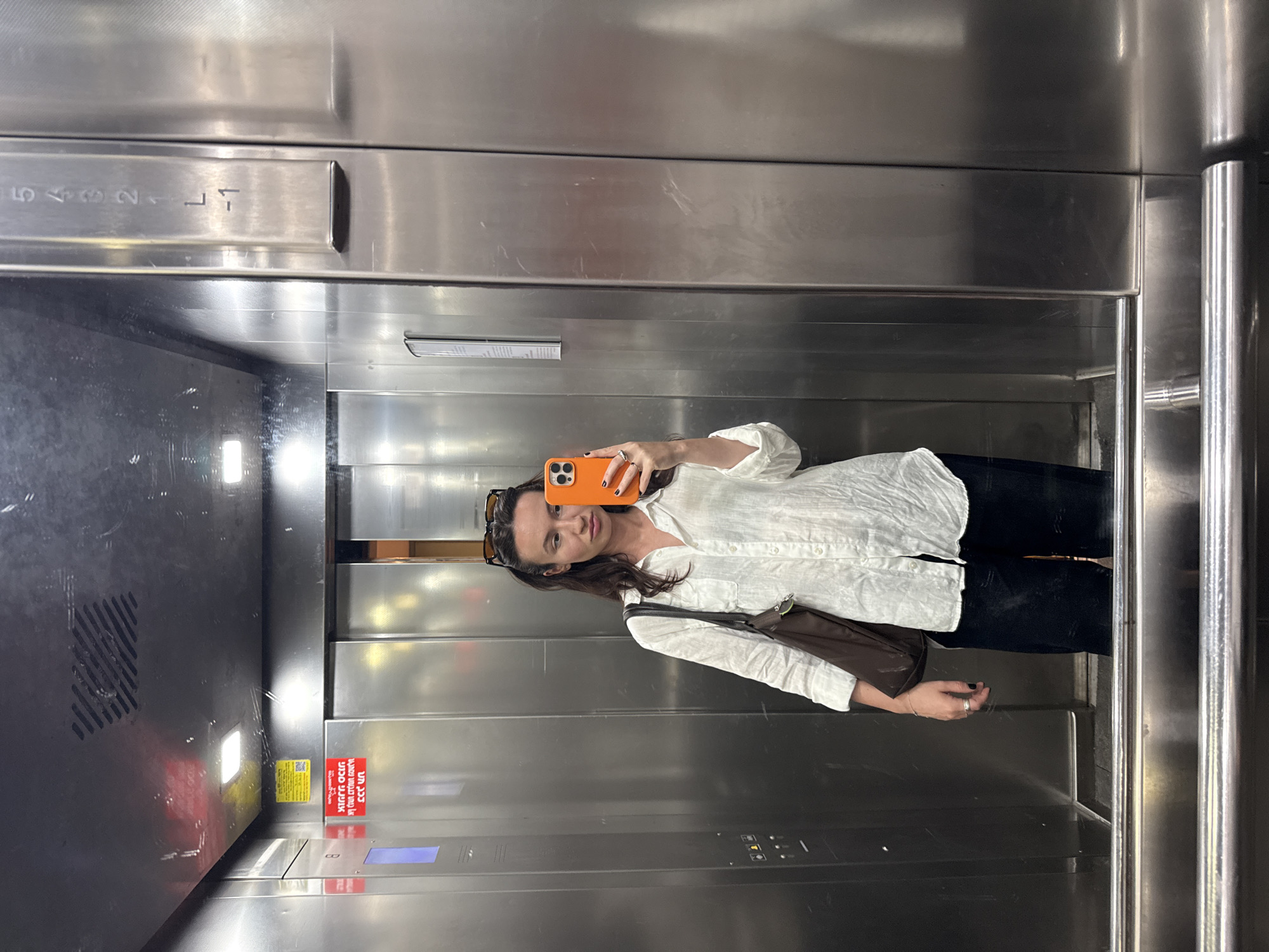 A woman is taking a selfie in an elevator. She is wearing a white button-down shirt and black pants. She has a brown bag over her shoulder and sunglasses on top of her head. She is holding an orange phone. The elevator has buttons labeled -1 to 5, and a notice written in Hebrew. The location is likely in Tel-Aviv, Israel, based on the Hebrew writing. The interior of the elevator is stainless steel.