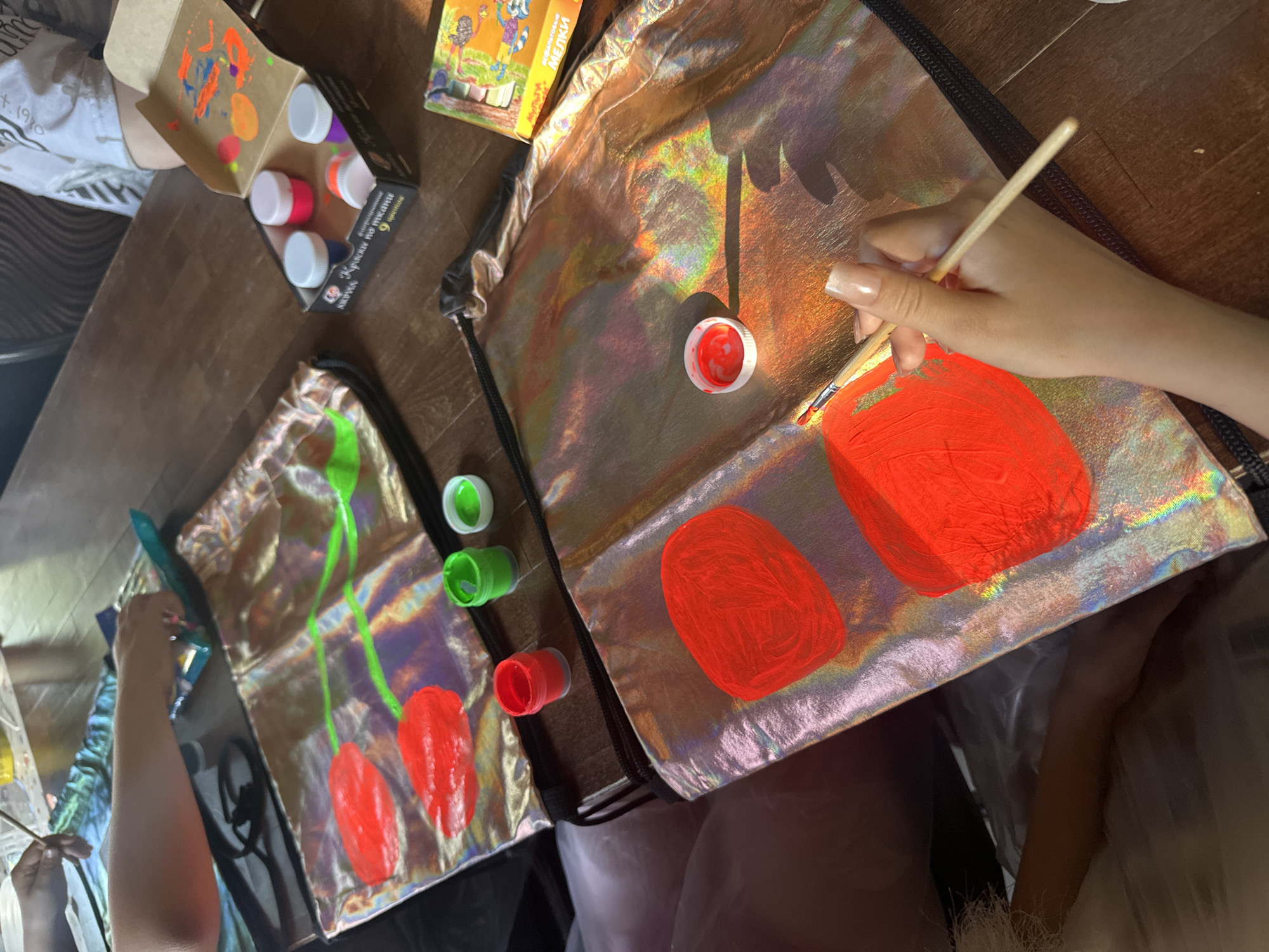 A close-up shot captures the artistic activity taking place on a wooden surface in the city of Unknown, Unknown. The scene focuses on two shiny drawstring bags laid out flat, presumably for painting. One bag features red oval shapes with green stems painted on it, resembling fruits or berries, while the other bag has similar red shapes being painted by a hand holding a paintbrush. Several small containers of colorful paints are scattered around the bags, along with a box of crayons or colored pencils. The environment suggests an arts and crafts session, possibly indoors, with natural light enhancing the vibrancy of the colors and the shiny texture of the bags.