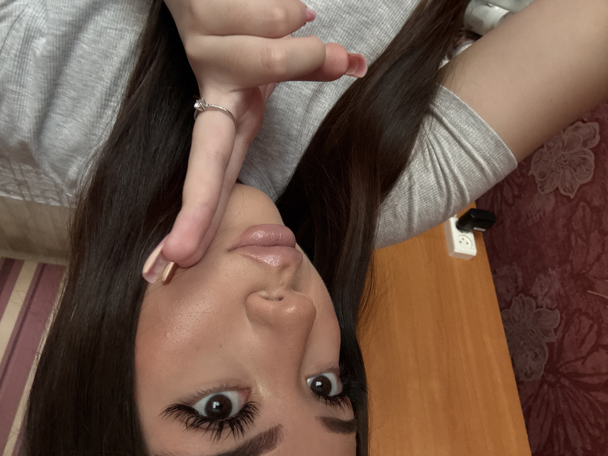 A close-up, rotated view of a woman's face. The woman has dark hair, brown eyes, and fair skin. She is wearing eyelash extensions and has a ring on her finger. The background includes a gray top, a wooden surface, an electrical outlet, and floral wallpaper. The image was taken indoors. There is no information in the image to indicate it was taken in Belopakhotnyi, Russia.