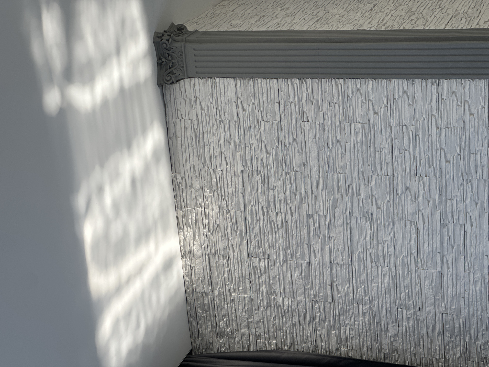 Here is a detailed description of the image:
The image shows an interior corner where two walls meet. The wall on the right is decorated with a textured, light gray material resembling stacked stone or tile with a vertical linear pattern. A darker gray decorative molding runs along the top of this textured wall, bordering the ceiling, which appears to have a subtly rough texture. The wall on the left is painted white and illuminated by soft, dappled sunlight creating abstract patterns of light and shadow. The base of the textured wall seems to meet a dark surface, possibly a floor or furniture. The scene is devoid of people or any clear indication of a specific location.