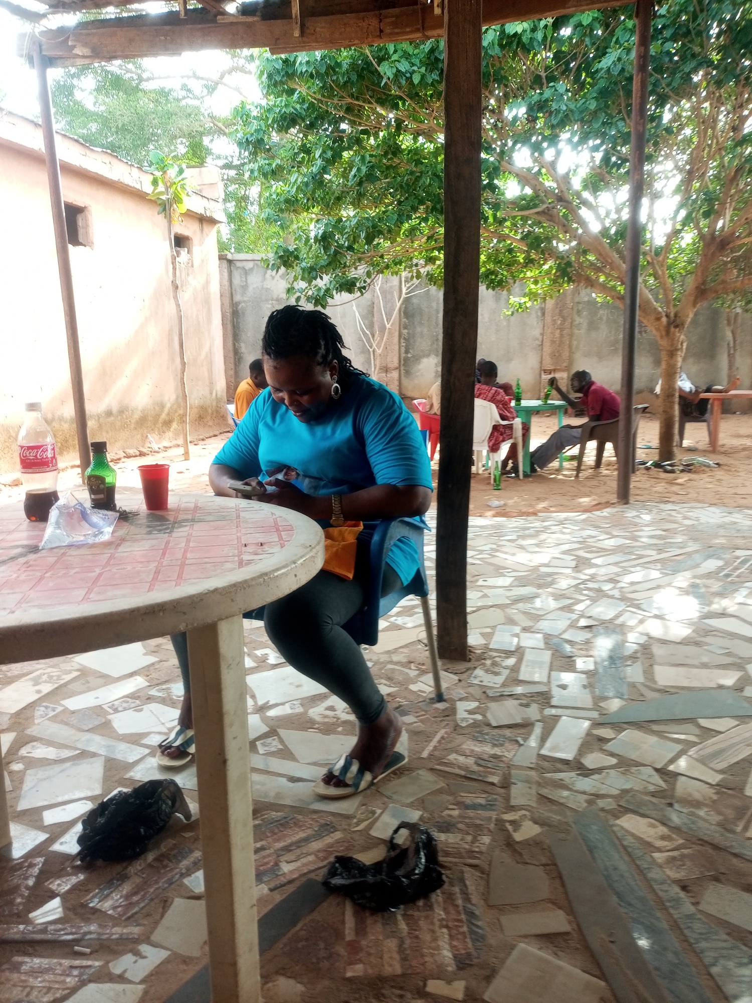 The image depicts a woman sitting at a round table outdoors, looking at her phone. The location appears to be a patio or open-air area, judging from the tiled floor and support beams. The city and specific place are unknown.
Other people are visible at tables in the background, and there are beverages on the table near the woman. The environment includes trees and a building in the background. There is debris/trash on the ground.