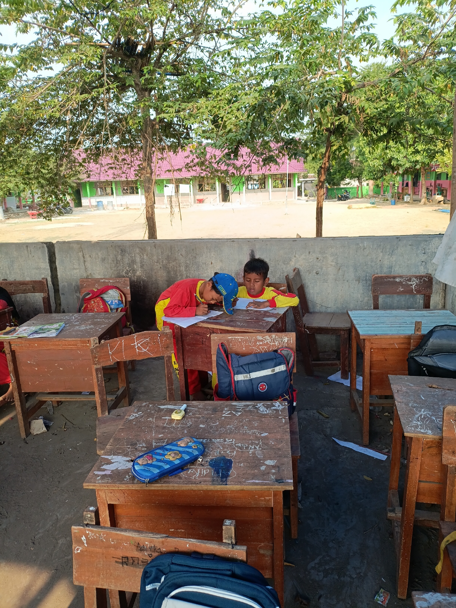 Two young boys, dressed in school uniforms, are seated at separate wooden desks outdoors. The boy on the right, wearing a yellow and red shirt and a blue cap, is leaning over his desk and writing on a piece of paper. The boy on the left, also in a yellow and red uniform, is similarly engaged with his work. The desks are old and worn, with visible graffiti. School buildings with pink roofs are visible in the background, set against a backdrop of trees and a clear sky. The ground beneath the desks appears to be dirt or sand. A blue pencil case with cartoon characters and a small yellow object, possibly a timer or a watch, are on one of the desks. Backpacks are also present, one with a Spider-Man logo. The scene appears to be a schoolyard in Sumberejo, Indonesia.