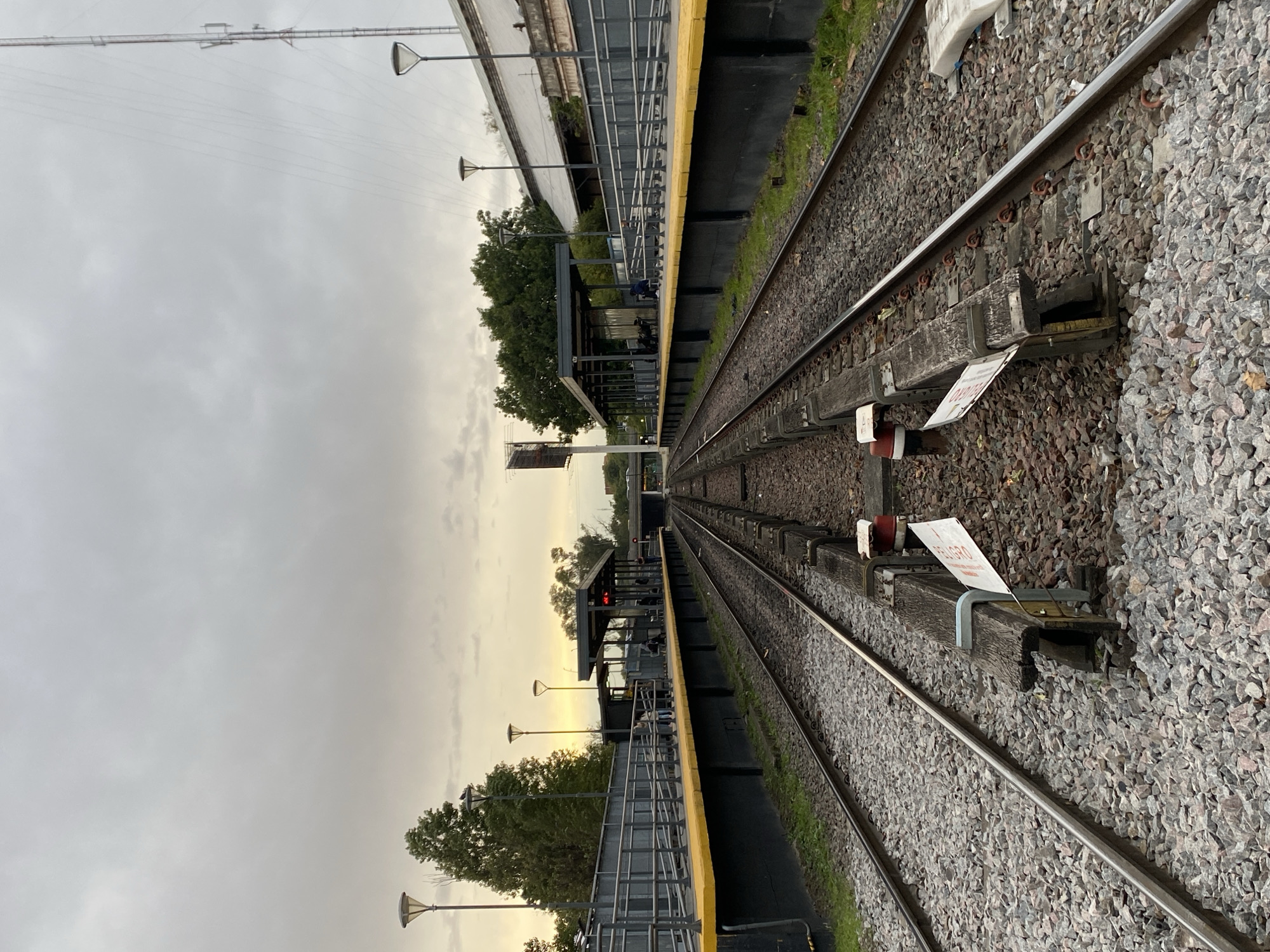 The image displays a train station in Sáenz Peña, Argentina, featuring parallel railway tracks and platforms under an overcast sky. The photograph is taken from near track level, looking down the length of the tracks as they recede into the distance.
On the right side of the image, two sets of railway tracks are clearly visible, laid on a bed of grey gravel ballast and wooden sleepers. A third rail or similar component runs parallel to the main rails. Attached to one of the wooden sleepers is a small white sign with red lettering, partially obscured, that reads "PELIGRO" (Spanish for "DANGER").
To the left of the tracks, a long platform stretches into the background, distinguished by a bright yellow safety strip along its edge and a metal railing. Further along this platform, there is a covered waiting area or shelter where indistinct figures of people can be seen. Underneath the shelter, a red digital display is visible, likely indicating train information. Several lamp posts with uniquely shaped lights are spaced along the platform. Trees and other foliage grow alongside the platform and in the background. In the far distance, additional station structures and a tall communication or power tower are visible against the sky.
The sky is predominantly overcast, but a warm, yellowish glow emanates from the horizon in the distance, suggesting either early morning or late afternoon light. The overall lighting is soft and diffused. The scene depicts a quiet, functional train station during a transitional period of the day.