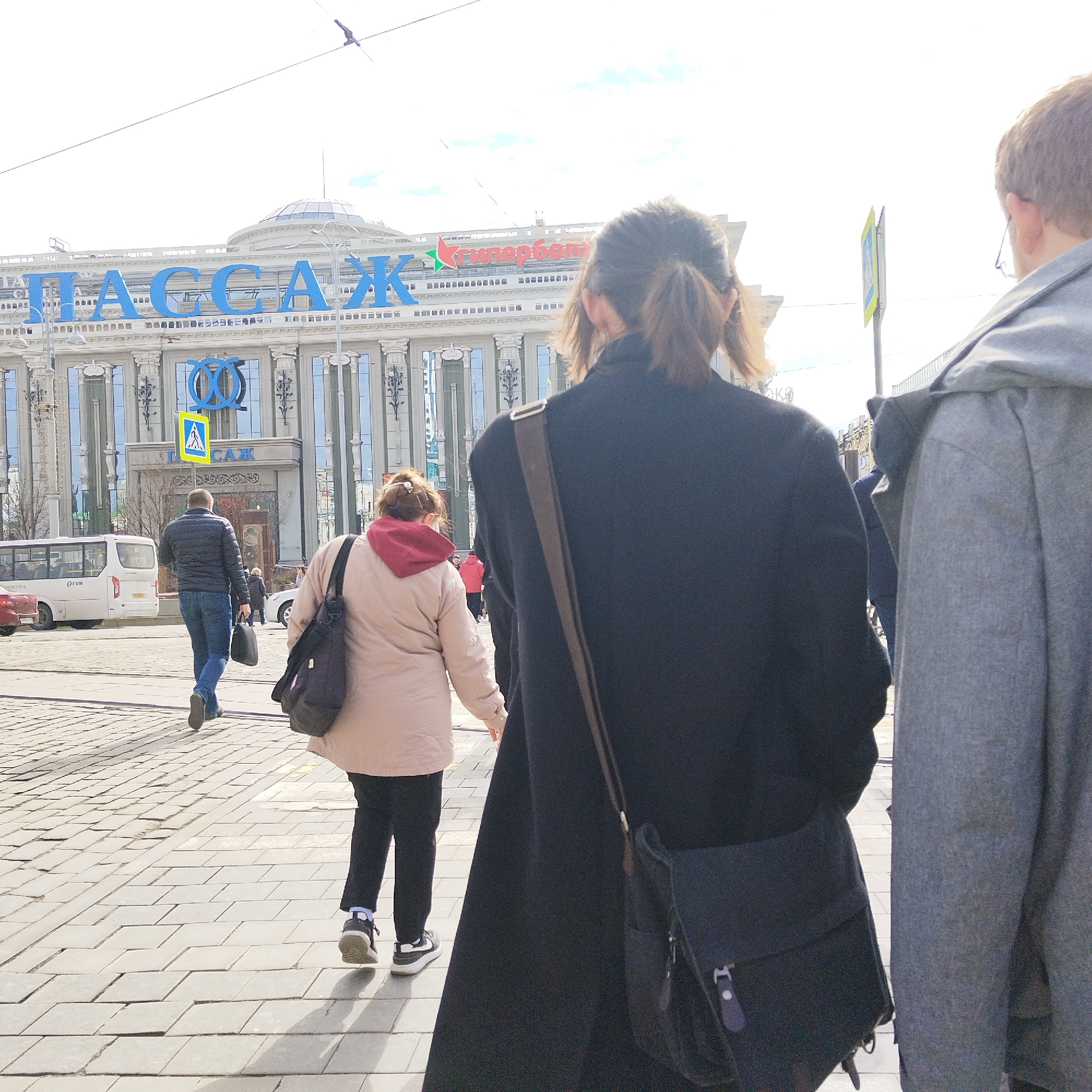 The image captures a street scene in Yekaterinburg, Russia. The architectural landmark "Passazh" with a prominent blue sign in Russian Cyrillic is visible in the background. People are walking on a pedestrian-paved surface, including a man walking away from the camera, a woman with a light-colored jacket and dark pants, and two people closer to the camera, one with a dark coat and bag and the other with a grey coat. A small bus is parked nearby. A pedestrian crossing sign is visible near the "Passazh" building. The sky is overcast, creating a bright daylight environment.