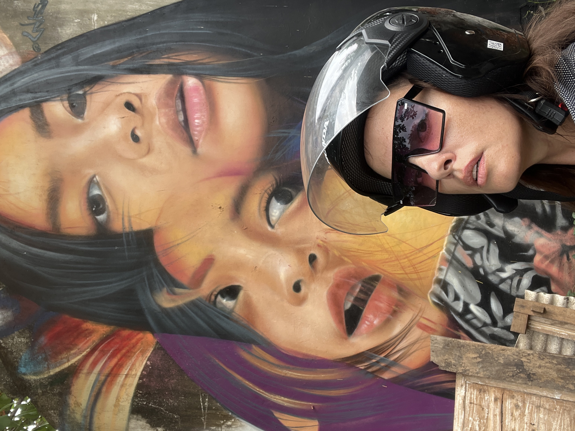 A woman wearing a black motorcycle helmet and oversized sunglasses is posing in front of a vibrant mural on a wall in Kuta, Indonesia. The mural depicts two young girls, rendered in a realistic style with bold colors. The woman's face is turned upwards, looking towards the mural, with her lips slightly parted. The helmet has a clear visor, reflecting the surrounding environment. In the foreground, a stack of weathered wooden planks and corrugated roofing material is visible, suggesting an urban or possibly semi-constructed area. The overall scene is colorful and striking, with the woman's presence adding a human element to the artistic backdrop.