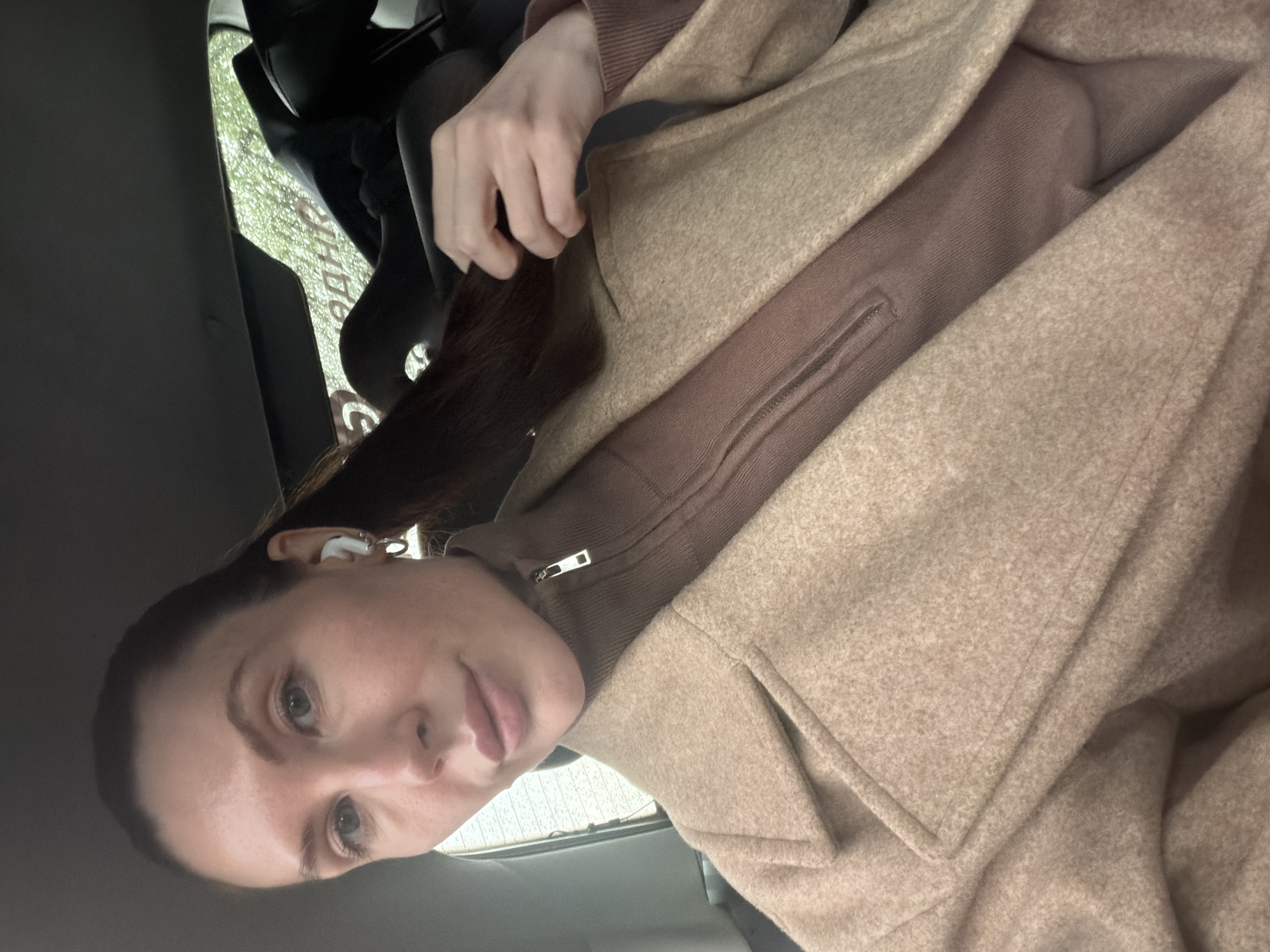 The image appears to be a selfie taken inside a car. A woman with fair skin and brown hair is the primary subject. She is wearing a brown turtleneck sweater and a beige overcoat. A portion of a car seat and a window are visible in the background. Based on the reflections visible through the glass, the location appears to be in Perm, Russia.