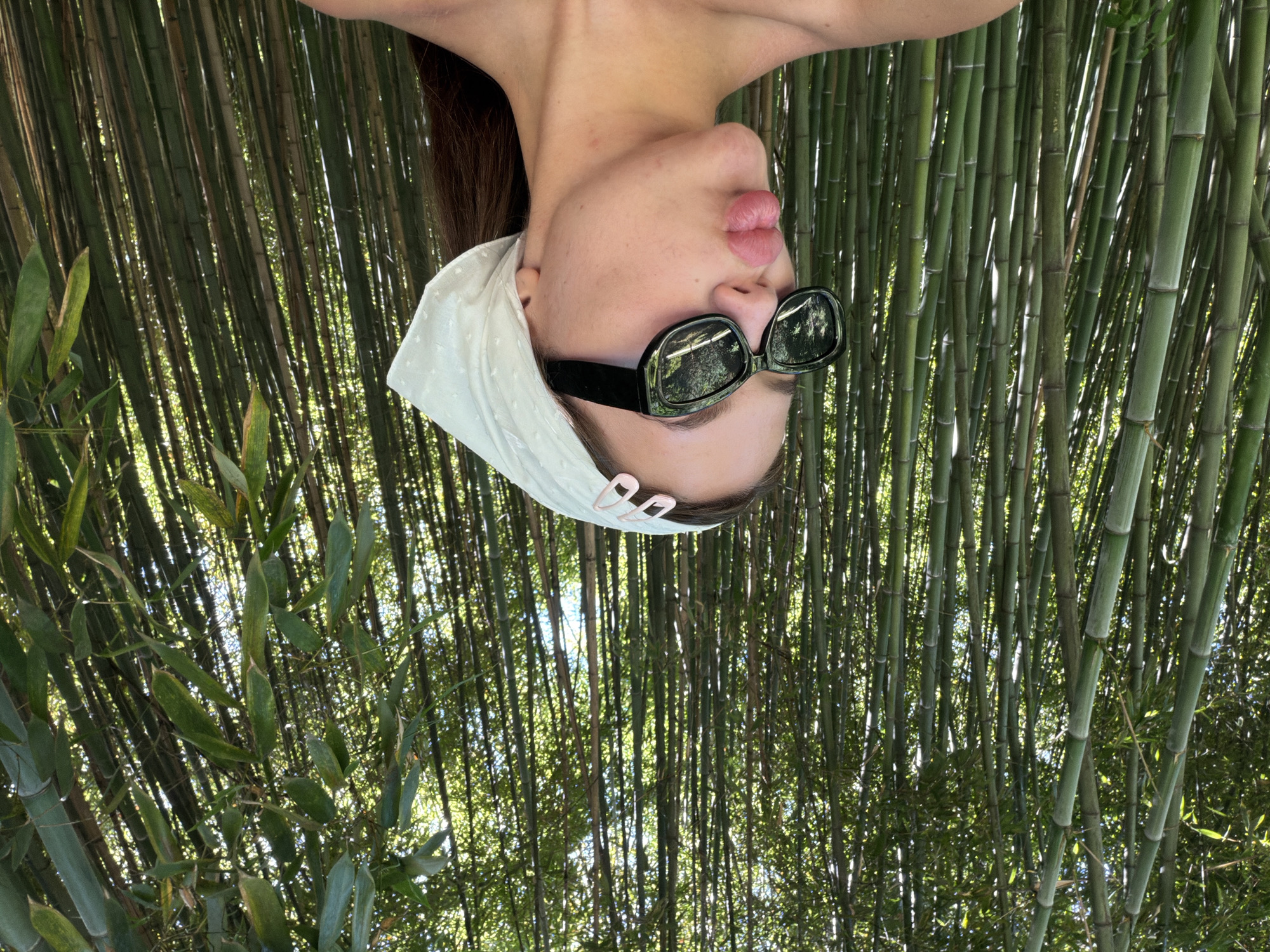 A young woman with brown hair is shown in a medium shot, posing for a photo in front of a dense bamboo forest. She is wearing black sunglasses and a white patterned bandana tied around her head, secured with two pink hair clips. Her lips are pursed, and she is looking slightly off-camera. The background is filled with tall, green bamboo stalks and leaves, with sunlight filtering through the foliage. The location is identified as Sirius, Russia.