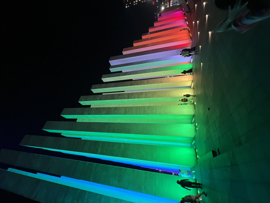 The image depicts a nighttime scene at Vinhomes Central Park, adjacent to the Landmark 81 building in Ho Chi Minh City, Vietnam. The central feature is a public art installation comprising approximately twenty-five rectangular, vertical pillars of varying heights, arranged in a diagonal line that slopes downwards from left to right, creating a staggered profile against the dark sky. Each pillar is individually illuminated from its base by colored LED lights, forming a continuous spectrum across the installation: transitioning from blue and cyan on the left, through various shades of green, yellow, and orange, to red and magenta on the right.
Approximately 10-15 individuals are present on the paved ground in front of and between the illuminated pillars. These individuals, mostly adults and some children, are seen walking or standing, observing the colorful display. On the far left, two figures walk towards the right. In the mid-left, a group of three, including a child, stands near the green-lit pillars. A solitary figure is silhouetted against an orange pillar, and a small cluster of three to four people gathers near the red-hued pillars on the mid-right. Part of a person's arm and leg are visible in the immediate lower-right foreground, suggesting someone seated nearby. The ground is a flat, paved surface, reflecting the colored light, and small recessed ground lights are visible on the right side. The background features a dark night sky with faint city lights and distant building outlines on the far right.