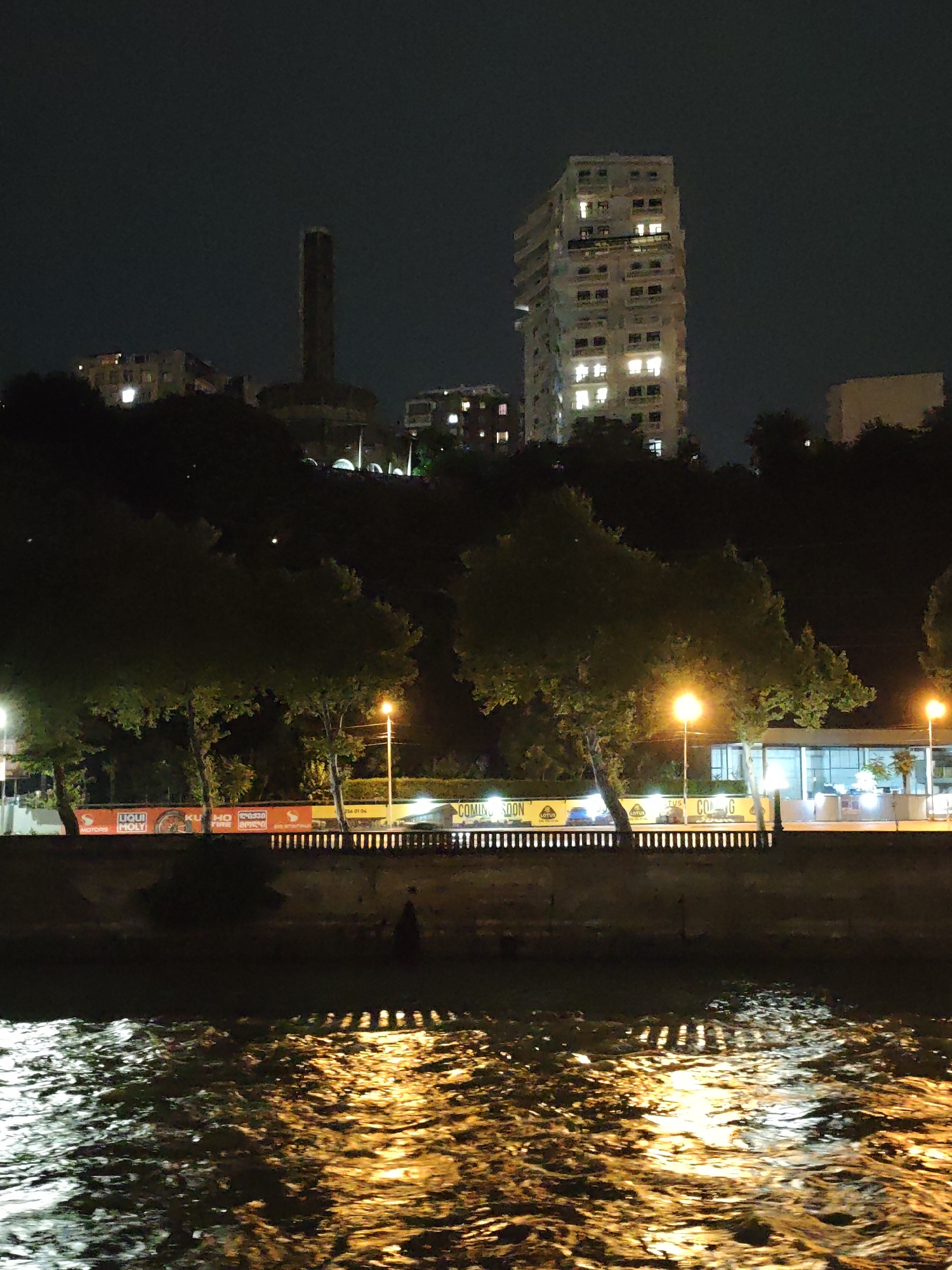 This is a nighttime photograph of the Mtkvari River in Tbilisi, Georgia. The scene is illuminated by artificial lights from the city and the reflections on the water's surface. On the far bank, a hill is covered with trees and buildings. A tall, slender, dark structure, possibly a chimney or monument, stands prominently on the left side of the hill. To the right of this structure, a modern, multi-story building with many lit windows is visible. In the foreground, a person is briefly visible standing near the water's edge. The river water is choppy, with strong reflections of yellow and white lights creating a dynamic pattern on its surface. Along the embankment, a low wall topped with a railing separates the river from a road or walkway. A banner displaying text including "LIQUI MOLY," "KUHO TIRE," and "COMING SOON" is stretched across this area. Streetlights are also visible along the embankment. The overall atmosphere is urban and nocturnal, with a sense of the city coming alive after dark.