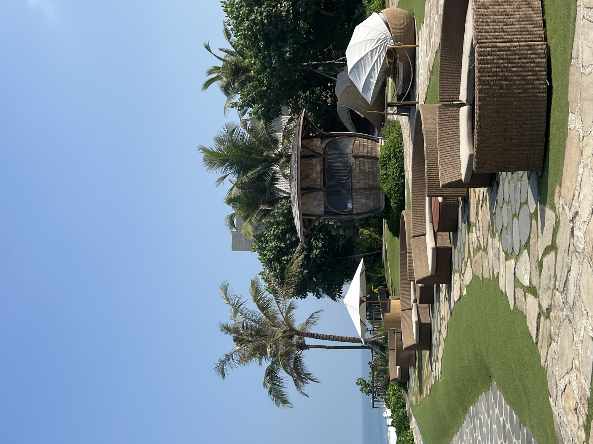 This image depicts an outdoor seating area with a coastal view. The scene is set in the city of Kediri, Indonesia, likely at a resort or restaurant by the sea. In the foreground, several circular rattan sofas with beige cushions are arranged on a manicured lawn, interspersed with a stone pathway. Behind the seating area, a peculiar wooden structure with a curved roof and large windows resembles a hut or pavilion. Further back, lush green trees and palm trees are visible, with a glimpse of the ocean and a white sandy beach in the distance. A white umbrella shades a seating area near the wooden structure, and a covered seating pod is also present. The sky is clear and blue, indicating a sunny day. There are no people or animals visible in the image.