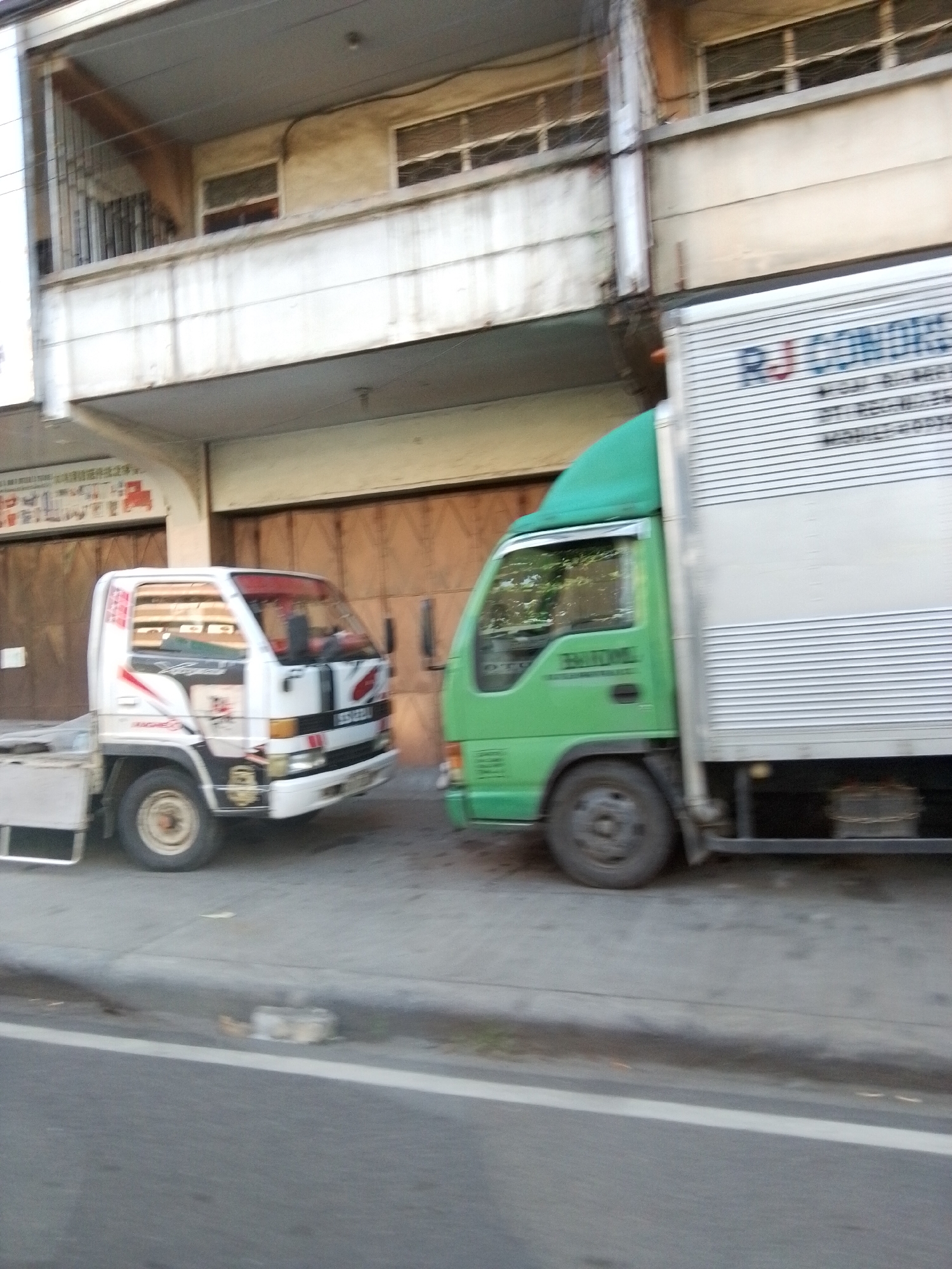 The image displays two parked trucks on a street in Davao City, Philippines, positioned alongside a multi-story building under daylight conditions. On the left, a white flatbed truck, likely an Isuzu, features red and black graphical accents on its cab and is oriented towards the right side of the frame. Adjacent to it on the right is a green box truck, also facing right, equipped with a silver, horizontally ribbed cargo box. The side of the green truck's box body exhibits partially legible text, including the characters "RJ LOR." Both vehicles show evidence of operational use. The multi-story building directly abuts the sidewalk, featuring large, closed doors at ground level and a prominent first-floor balcony with a metal railing. Two windows are visible on the first floor above the ground level. The building's light-colored exterior shows signs of weathering. A concrete sidewalk separates the parked trucks from the building, and an asphalt roadway with a solid white lane marker is visible in the foreground. No individuals are present in the scene.