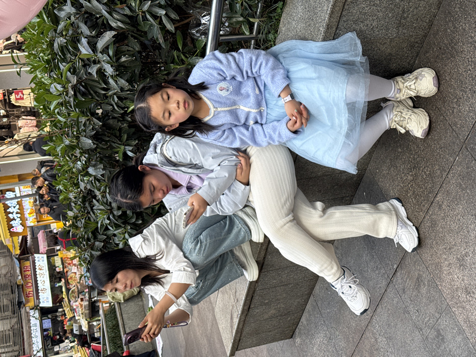 The image captures three East Asian females situated on a stone ledge in an outdoor commercial street environment within Jiangjin, China. On the left, an adult female in a white shirt and sparkling light-blue pants is crouched on the ledge, actively viewing a dark-colored smartphone held in both hands. Adjacent to her, a female teenager or young adult, dressed in a light-gray jacket over a purple top and cream-colored ribbed pants, is seated and engaged with a white smartphone. To her right, a young girl, approximately 5-8 years old, sits upright. She wears a light-blue cardigan with an 'Elsa' emblem, a light-blue layered skirt, white tights, and a white digital watch on her left wrist; her hands are clasped, and her gaze is directed slightly downwards. A dense green hedge is positioned behind the individuals. The immediate foreground shows grey tiled pavement. The background reveals a bustling urban street with various food stalls and shops, identifiable by Chinese signage such as '伤心凉粉' and '辣妹子火锅', displaying goods and populated by multiple pedestrians. No direct interaction among the three individuals is depicted; the two older females are absorbed in their electronic devices, while the youngest is passively seated.