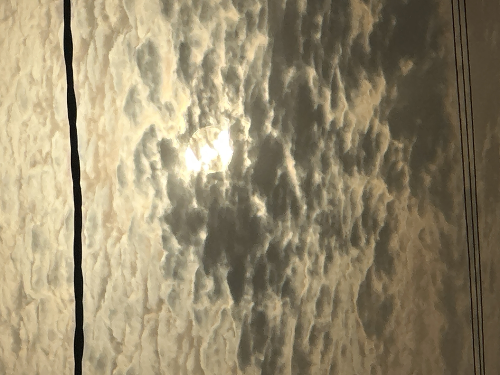 The image shows a cloudy sky with the sun shining through a gap in the clouds. Two sets of power lines are visible on the left and right sides of the image. The scene likely depicts a daytime view of the sky in Bukuru, Nigeria.