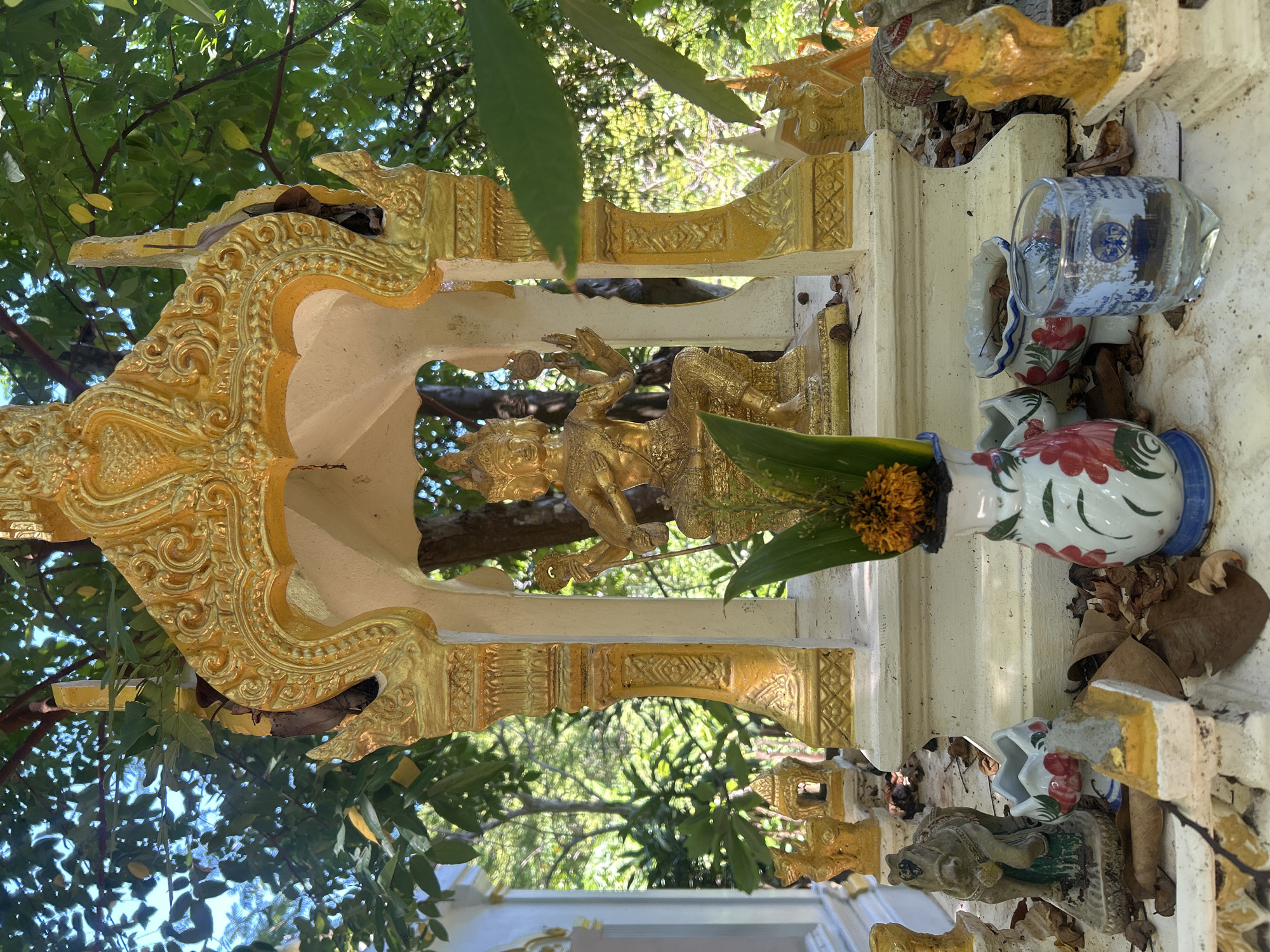 The image shows a small, ornate shrine outdoors, likely in Koh Phangan, Thailand. The shrine is topped with a golden, tiered roof structure with intricate carvings. Inside the shrine, a golden statue of a deity with multiple arms sits cross-legged. In front of the shrine, a white vase with floral patterns holds green leaves and a cluster of orange flowers. Scattered around the base of the shrine are fallen leaves, small decorative figurines of animals and architectural elements, and a glass containing a clear liquid. The background consists of lush green foliage and trees, suggesting a tropical environment. The overall scene depicts a religious offering or a place of worship in a natural setting.