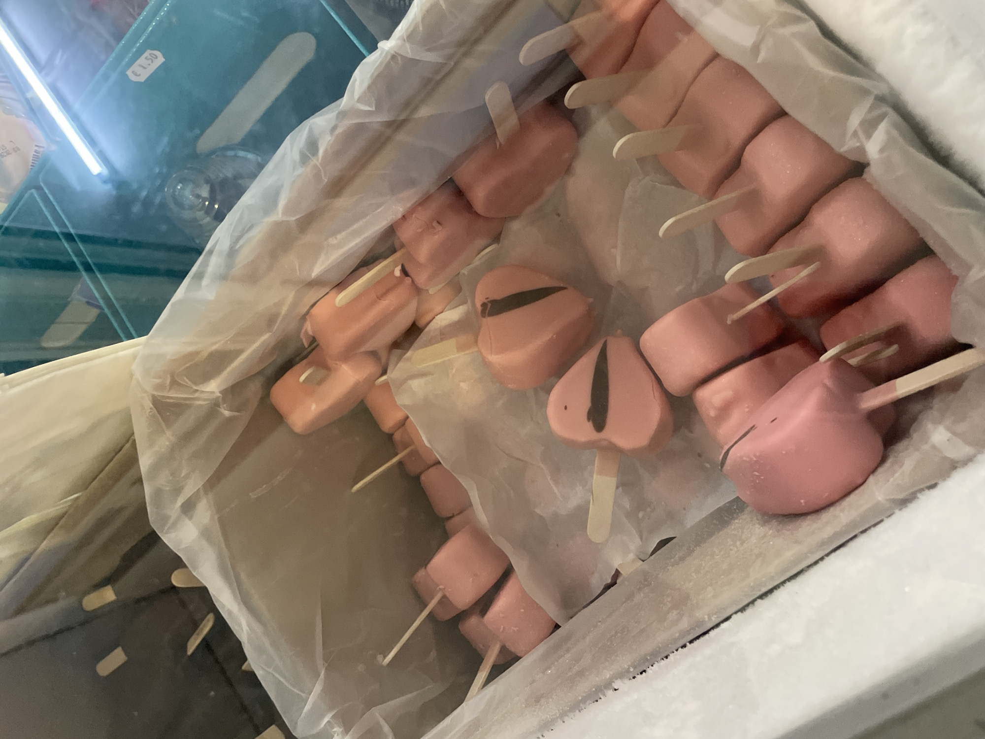 The image shows a close-up, high-angle view of a chest freezer containing numerous pink, heart-shaped and rectangular popsicles on sticks. The popsicles are arranged in a chaotic manner, some partially covered by white paper or plastic. The freezer is lined with white plastic. In the background, a glass display case is visible, with some items inside and a price tag of "€ 1.50." The scene appears to be an interior view of a commercial establishment, likely a shop or kiosk, possibly in Skala Avdiron, Greece, given the presence of Euros. There are no people visible in the image.