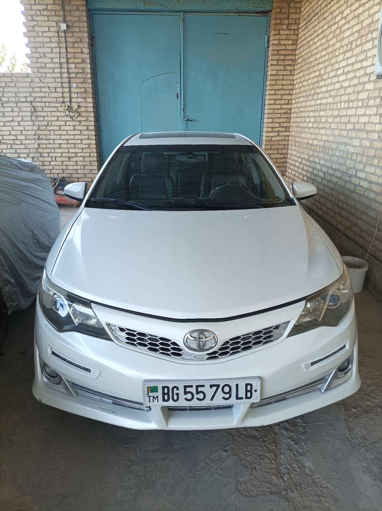A front-facing, eye-level, full shot captures a white 2012 Toyota Camry parked inside a garage in Seýdi, Turkmenistan. The vehicle is positioned centrally, with its front bumper and grille taking up a significant portion of the lower frame. The car's headlights are illuminated, and a license plate, "BG 5579 LB," is clearly visible on the front. A grey car cover is draped over an unseen vehicle to the left of the Camry, partially obscuring it from view. The background features a light-colored brick wall on the left and right sides, with a large, teal-colored metal door dominating the center. The floor is concrete, showing signs of wear and dirt. The overall lighting suggests an indoor, possibly dimly lit environment.