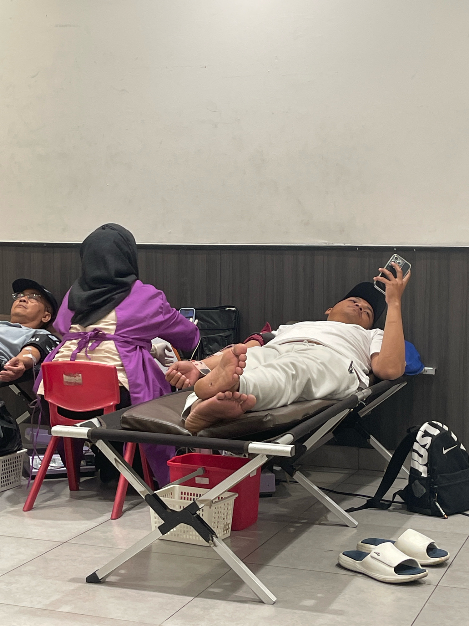 The image depicts a blood donation setting, possibly in a community hall or medical facility, located in Pagedangan, Indonesia. In the foreground, two individuals are lying on cots, undergoing or having completed blood donation. The person on the right, a young male wearing a white t-shirt and pants, is propped up, holding a smartphone and looking at it. His feet are visible, showing tubes connected to his arm. To his left, another cot is occupied by a person whose arm is also connected to medical equipment.
Behind the cots, a woman wearing a purple hijab and a matching long-sleeved garment, possibly a nurse or phlebotomist, is attending to the person on the left cot. She is wearing gloves and appears to be in the process of drawing blood. A red plastic chair is positioned between the two cots.
The environment is a clean, indoor space with a light-colored wall in the background. The floor is tiled with a grey pattern. Various objects are scattered around, including a white plastic basket, a red plastic bucket, and a black backpack with a white Nike logo. A pair of white slippers rests on the floor near the bottom right corner. The overall scene conveys a sense of a public health activity taking place in a functional setting.