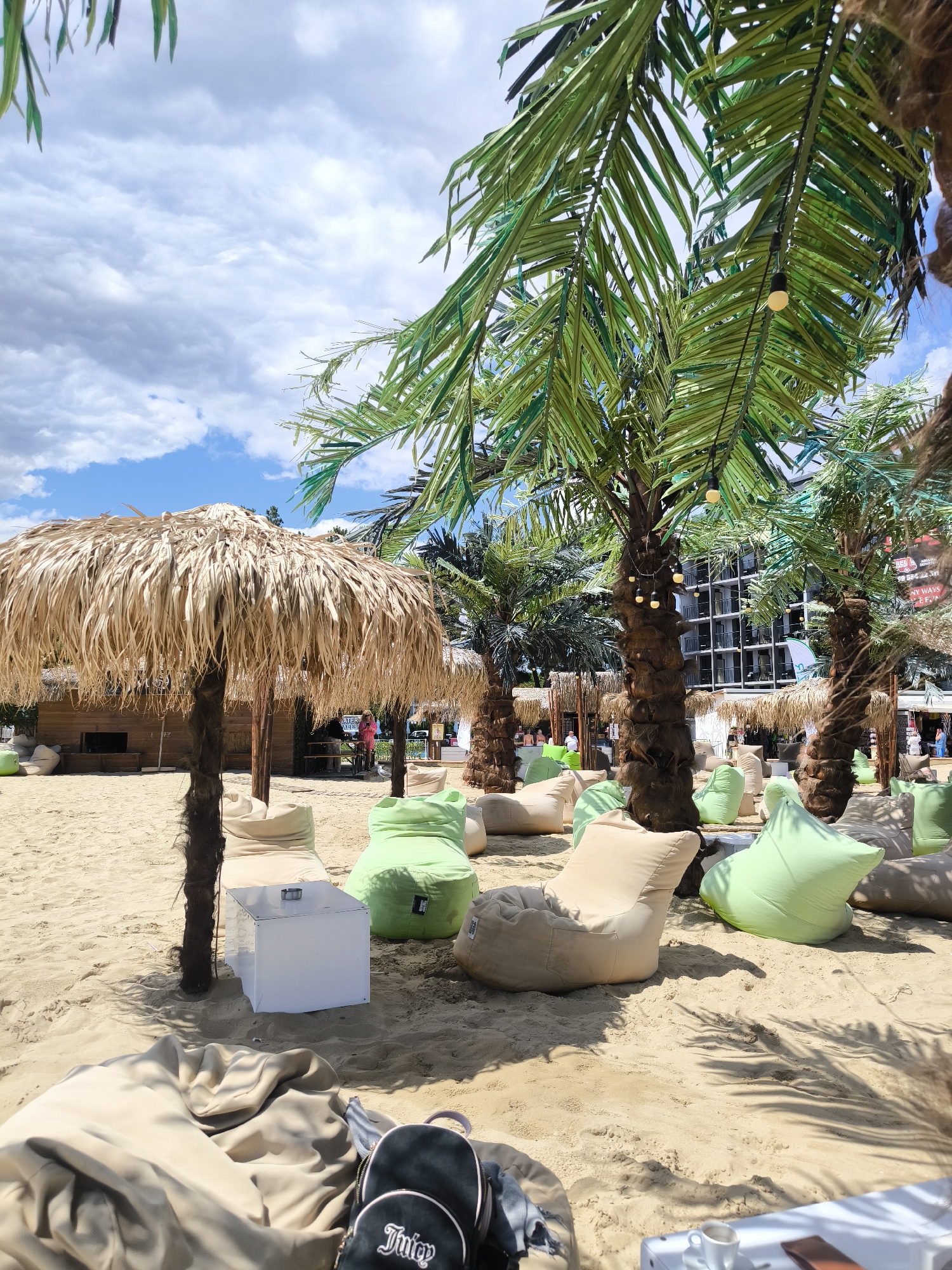 This image depicts an outdoor seating area on a sandy beach, likely a bar or lounge, in Nesebar, Bulgaria. The scene is dominated by several artificial palm trees with decorative lighting. Various beanbag chairs in light green and beige are scattered across the sand, suggesting a relaxed atmosphere. In the background, a wooden structure with a thatched roof, possibly a bar or kiosk, is visible, along with a modern building with multiple balconies. A few people are present in the background, some standing and others seated. A white cubic table with an ashtray sits near a thatched umbrella in the foreground, and a small backpack with the "Juicy" logo is visible on a beanbag chair. The sky is partly cloudy with blue patches.