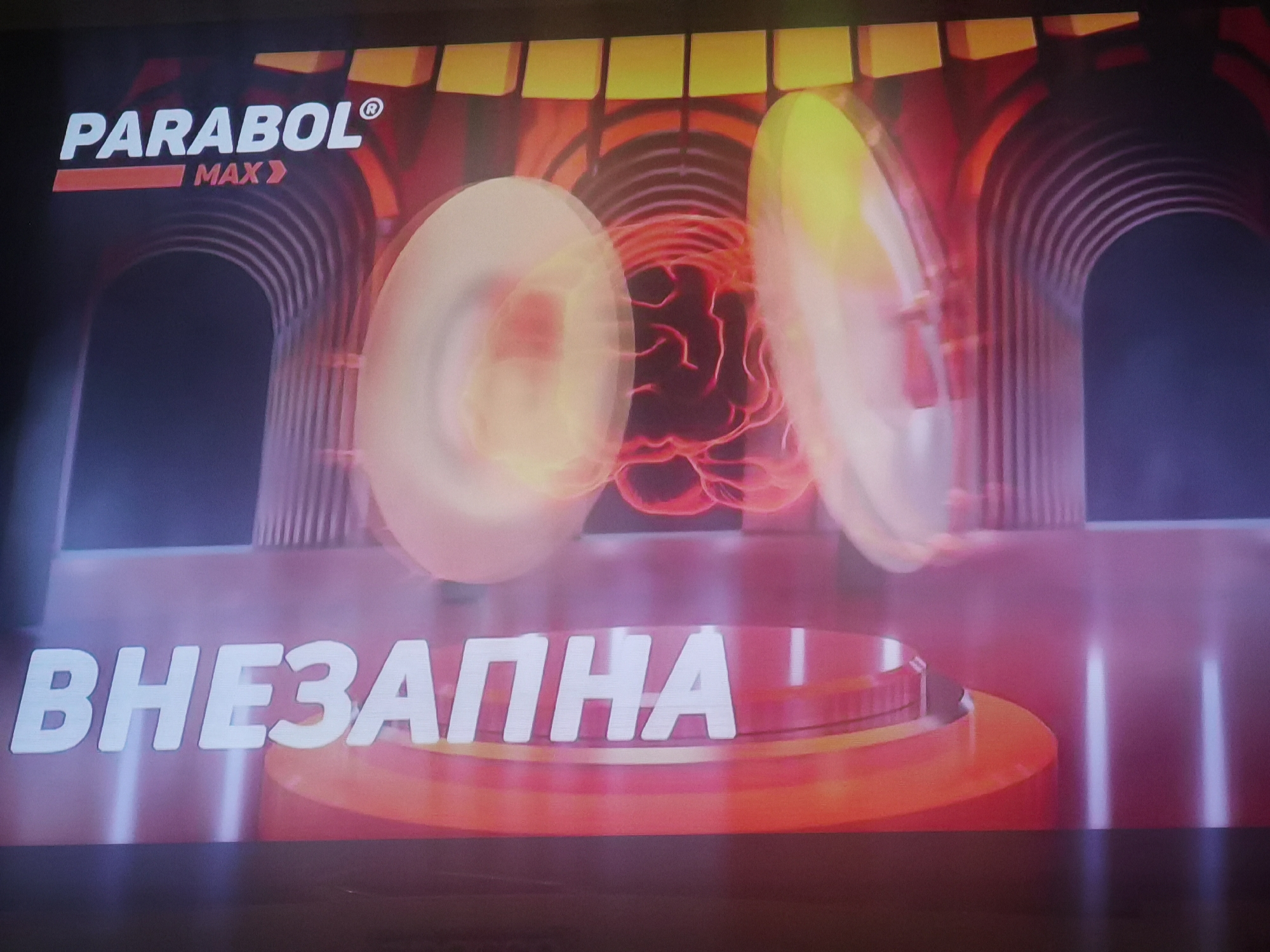 This image is a digital graphic, likely a screen capture, presented in a context related to the city of Lomets, Bulgaria. The upper left quadrant displays the white capitalized text "PARABOL®" with a registered trademark symbol, positioned above a red horizontal bar containing the white capitalized text "MAX»". The background features abstract architectural elements resembling a series of reddish-brown, arched tunnels receding into a dark, blue-hued distance. Above these arches, a horizontal row of yellow, rectangular blocks forms a border. Central to the composition are two large, blurred, translucent, oval-shaped objects with bright edges, flanking a faintly glowing, red, organic, brain-like structure positioned between them. In the foreground, large, white, three-dimensional Cyrillic text spells "ВНЕЗАПНА", which translates to "SUDDEN" or "SUDDENLY". This text rests on a tiered, circular platform rendered in shades of red and orange with a reflective surface. The lower portion of the image shows a dark, reflective floor with vertical light streaks, suggesting an abstract digital environment. No people or explicit actions are depicted.