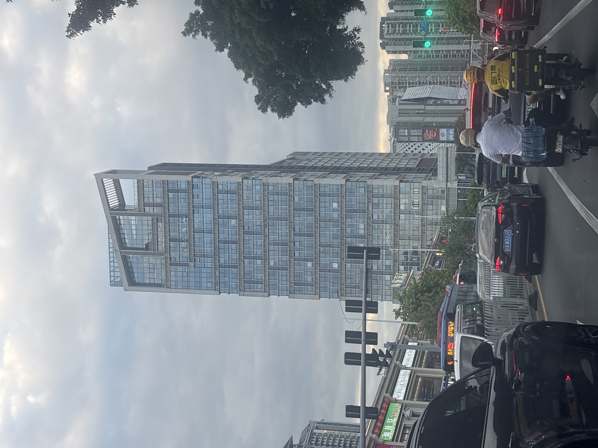 The image depicts a street scene in the city of Liwan District, China, under an overcast sky. A tall, modern skyscraper with a glass facade dominates the center of the frame. Traffic is visible on the road, including cars, a bus, and two individuals on electric scooters. The scooter riders are wearing helmets, and one is dressed in a white shirt, while the other wears a yellow shirt with text and a logo. The scooter behind the person in the yellow shirt has a large black storage container. Traffic lights are illuminated, showing green signals. Buildings, some with contemporary architectural designs, are seen in the background. Trees line the street, adding greenery to the urban environment.