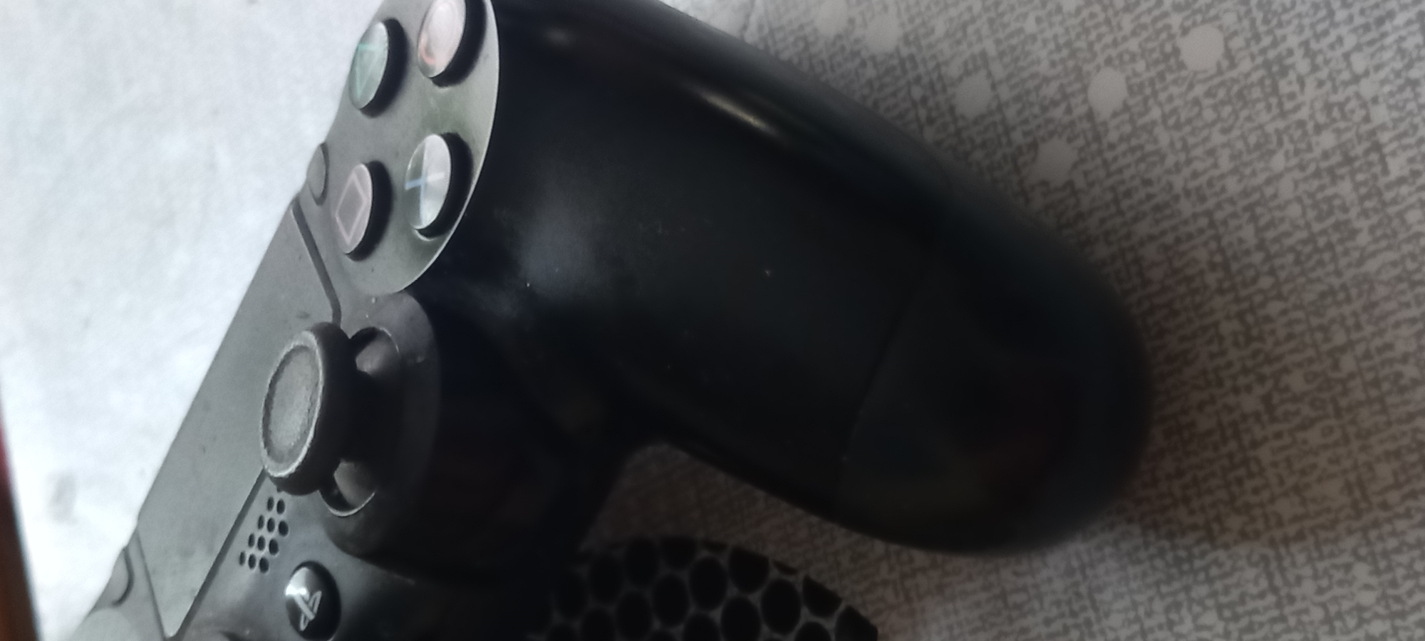 The image depicts a black Sony PlayStation 4 (PS4) DualShock 4 controller, positioned diagonally across a patterned surface. The controller is viewed from a slightly elevated angle, revealing its top surface, including the left analog stick, the PlayStation button, and the four face buttons (Triangle, Circle, Cross/X, and Square) on the right side. The left analog stick cap shows visible signs of wear and a textured, possibly deteriorated, surface. The controller's black plastic body exhibits minor surface marks and a used appearance, particularly on the right grip.
The controller rests on a light grey or off-white surface characterized by a subtle, repeating grid or interwoven linear pattern, interspersed with irregularly shaped lighter dots or blotches. Along the extreme left edge of the frame, a portion of a darker material with a distinct honeycomb or hexagonal pattern is partially visible beneath the controller.
The image provides no discernible visual information or contextual clues to determine its specific geographical location, including within the city of Kryvyi Rih, Ukraine.